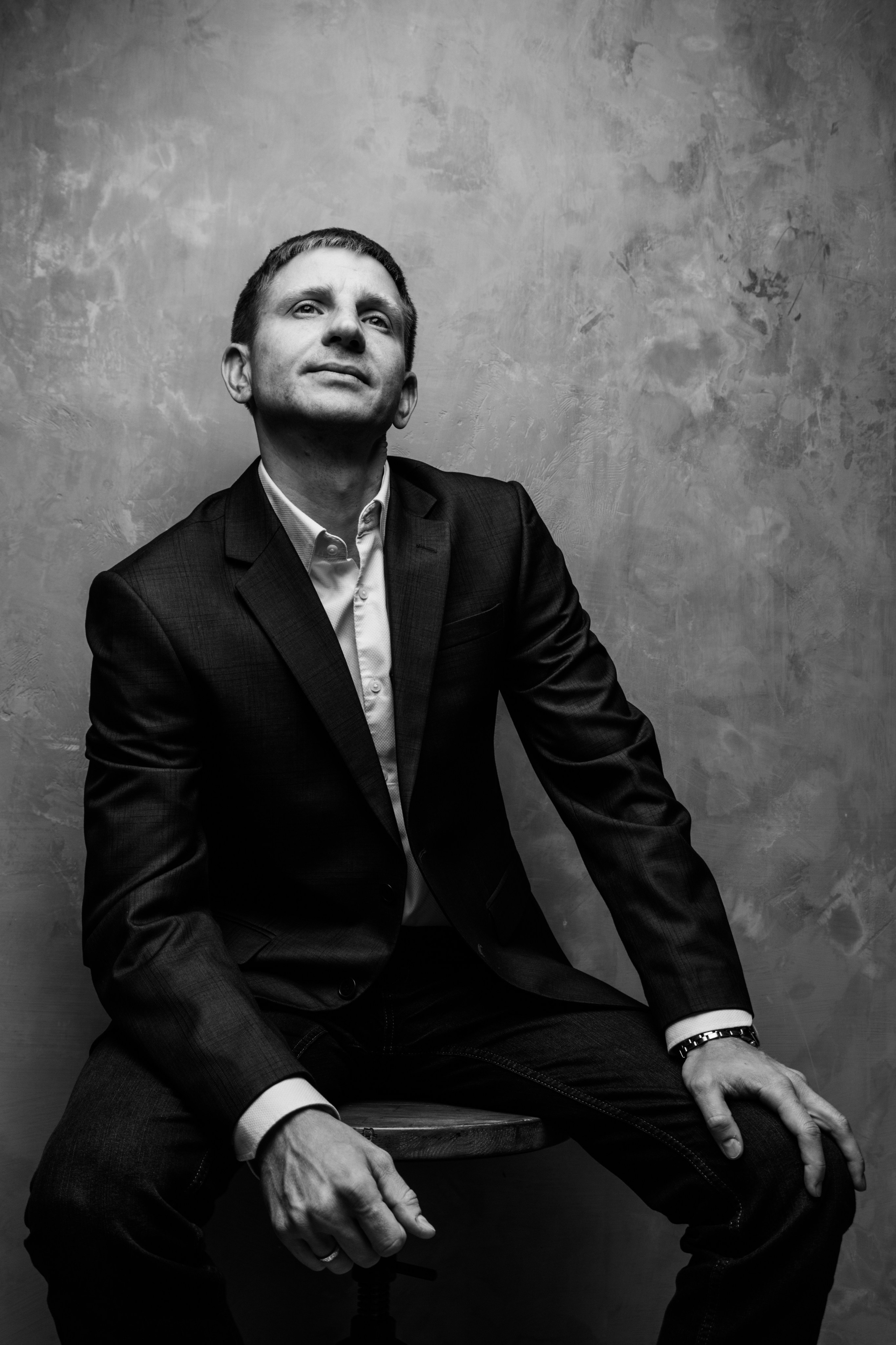 Черно-белый портрет мужчины в костюме на фактурном кирпичном фоне.