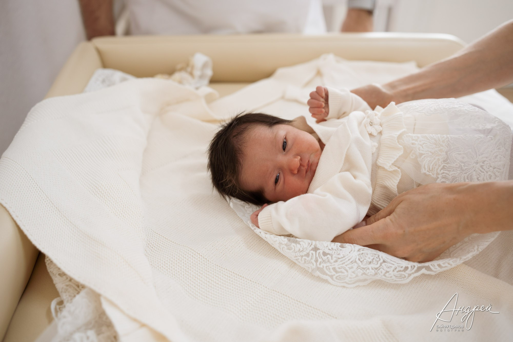 Новорожденный в белой одежде на кровати, его руки держат руки родителей — фотосъемка младенца в Ростове-на-Дону.