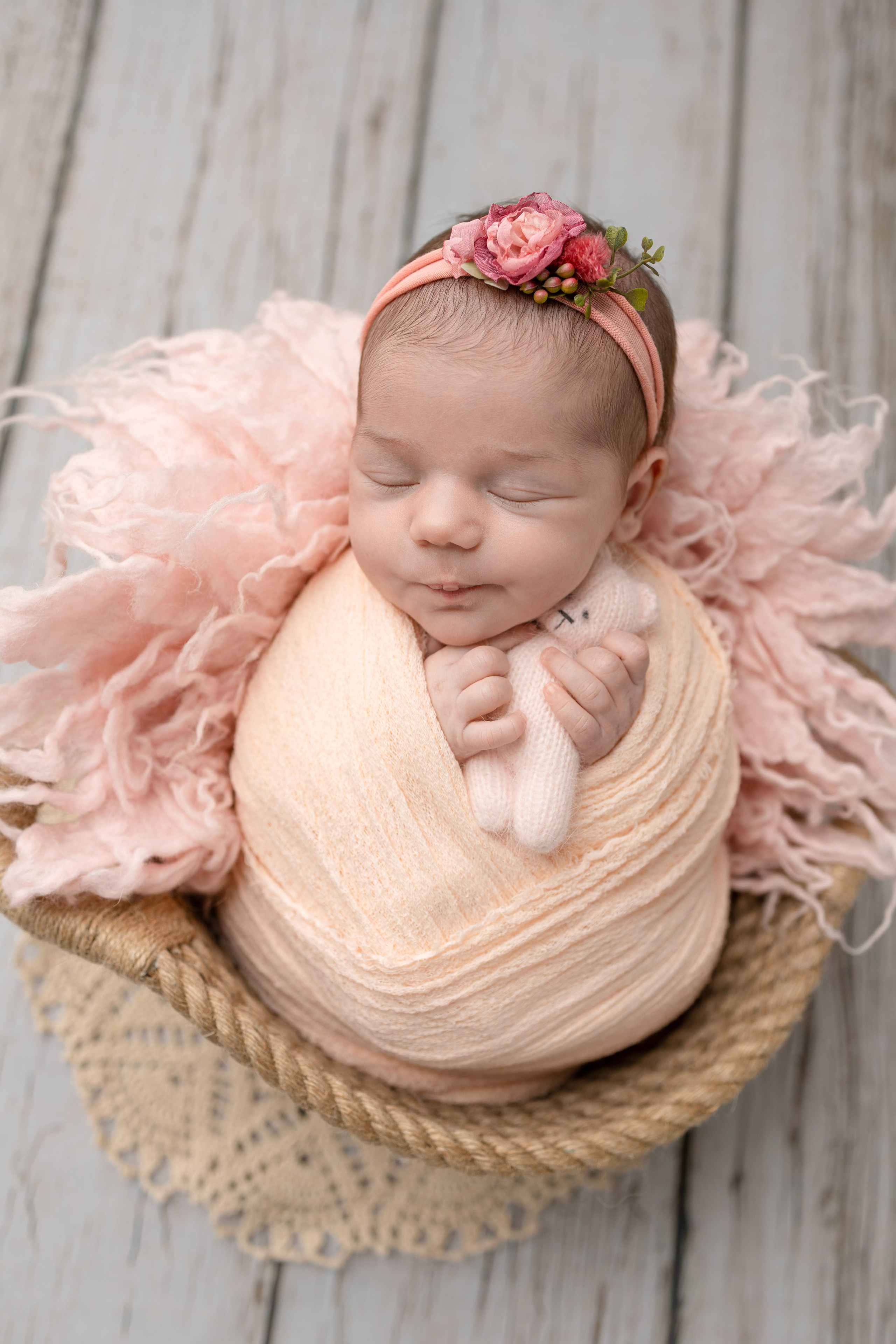 Новорожденный в розовом свертке с цветочным ободком, уложенный в корзине на фоне деревянной доски.