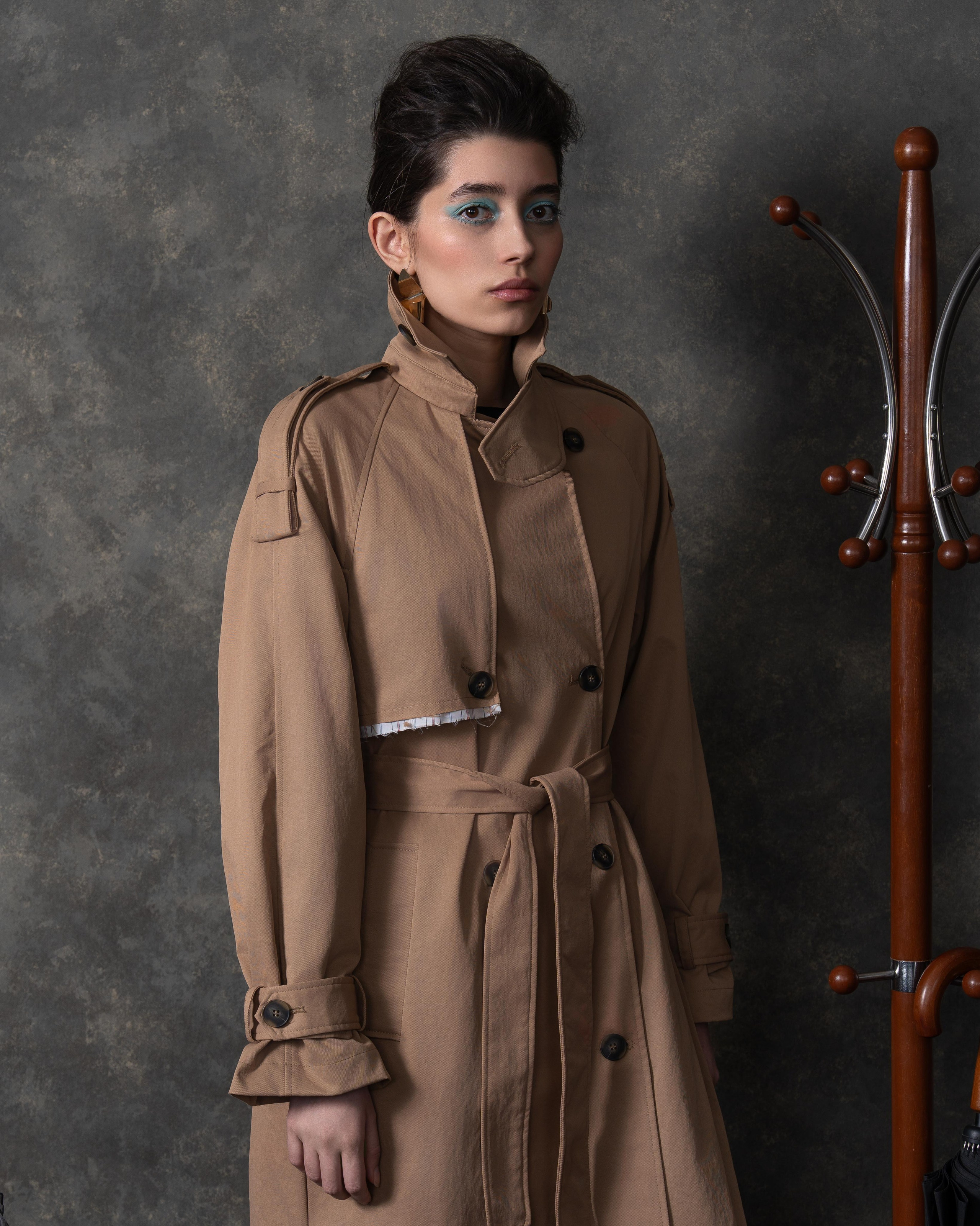 На фотографии изображена женщина, стоящая на фоне серой стены. Она одета в длинное бежевое пальто с поясом и большими пуговицами. Пальто имеет классический крой с воротником и карманами. Женщина смотрит прямо в камеру, её взгляд серьёзный и уверенный. У неё тёмные волосы, собранные в аккуратную причёску, и яркий макияж с акцентом на глаза, где видна синяя подводка. На её лице также заметны румяна и помада. В правой части кадра видна часть деревянной вешалки с крючками, на которой висит чёрный зонт. Освещение на фотографии мягкое, создающее тени на лице и одежде модели.
