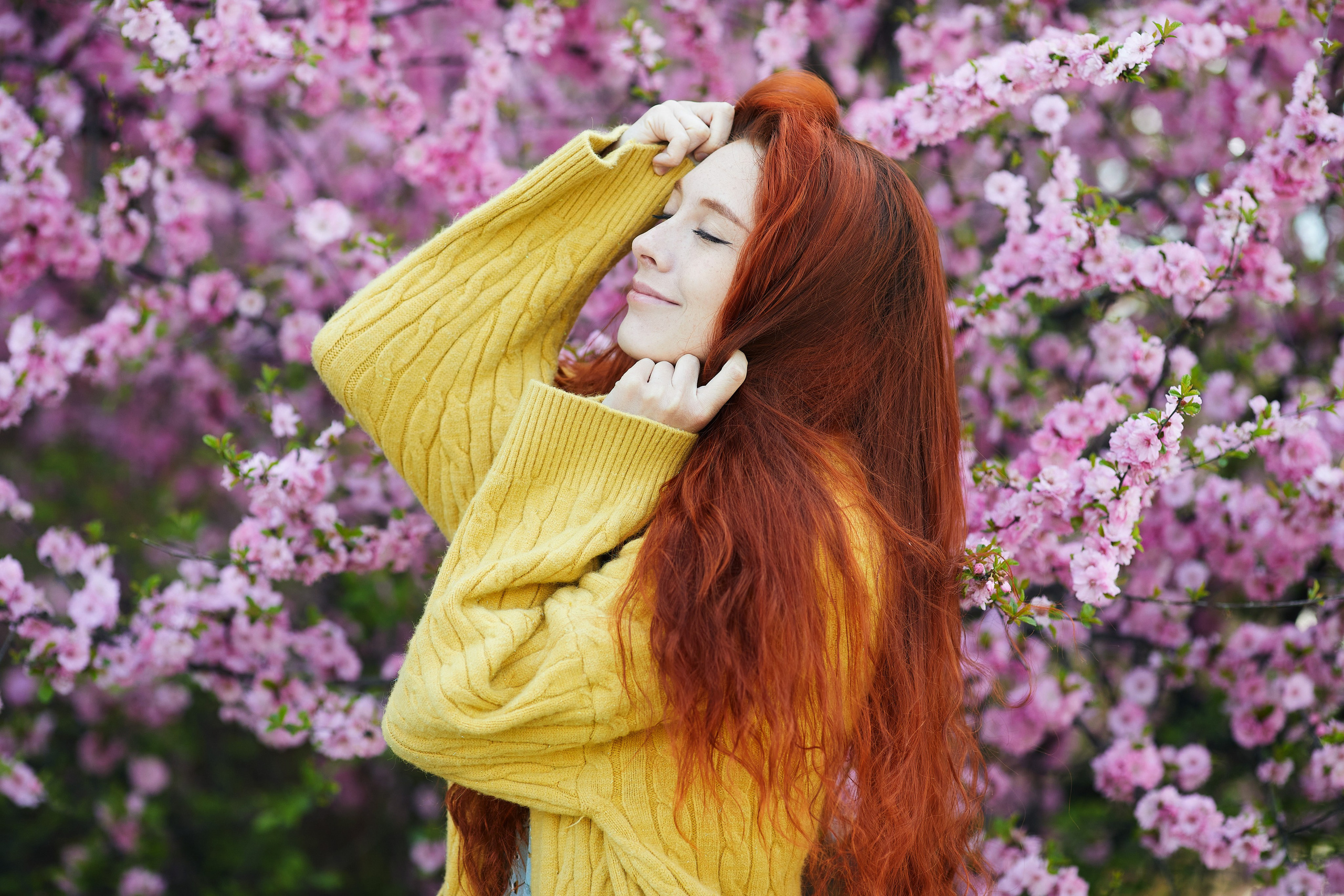Рыжеволосая девушка в тёплом жёлто-оранжевом свитере позирует на фоне цветущего куста в Находке