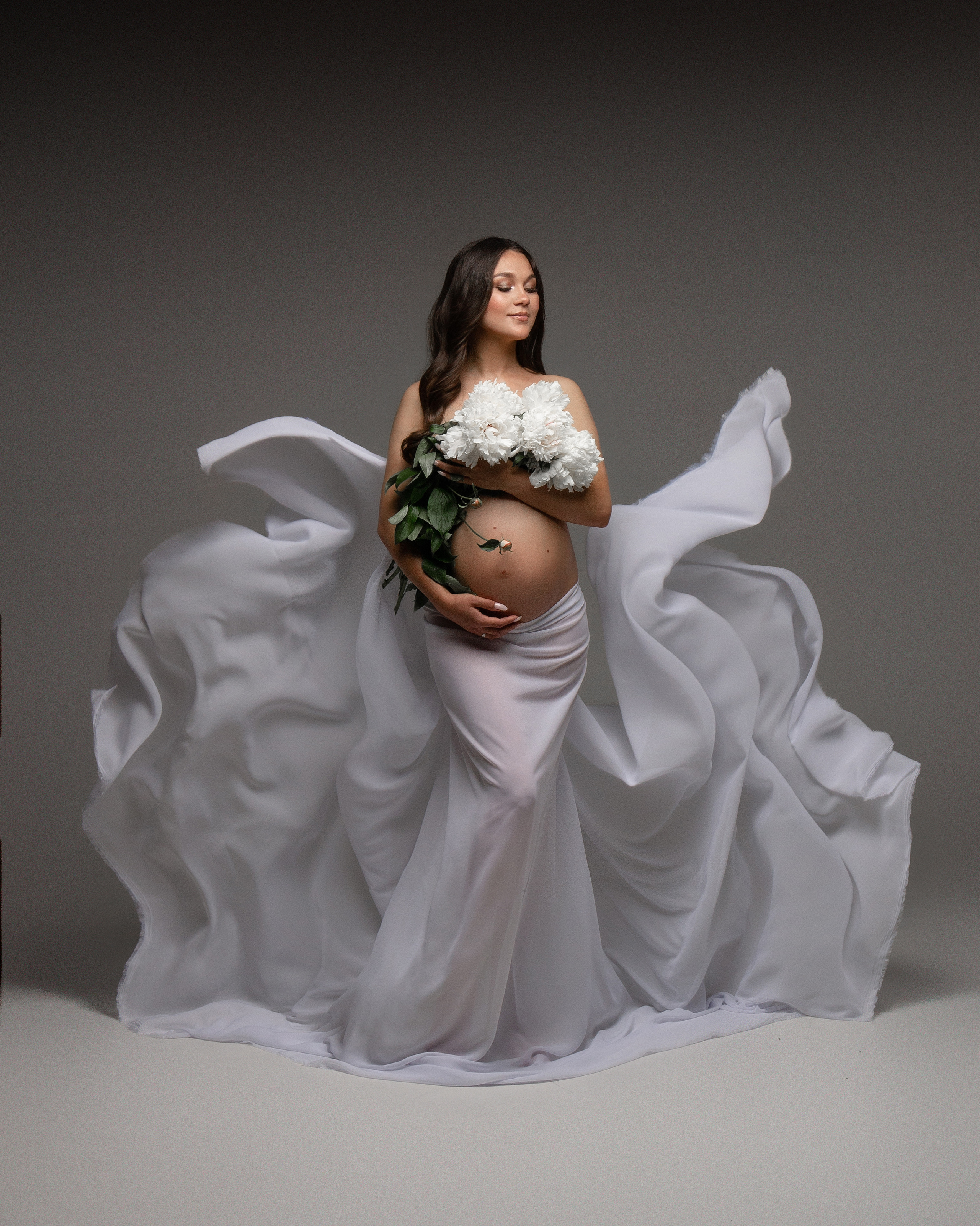 Беременная девушка с цветами в летящей белой ткани с пионами в руках