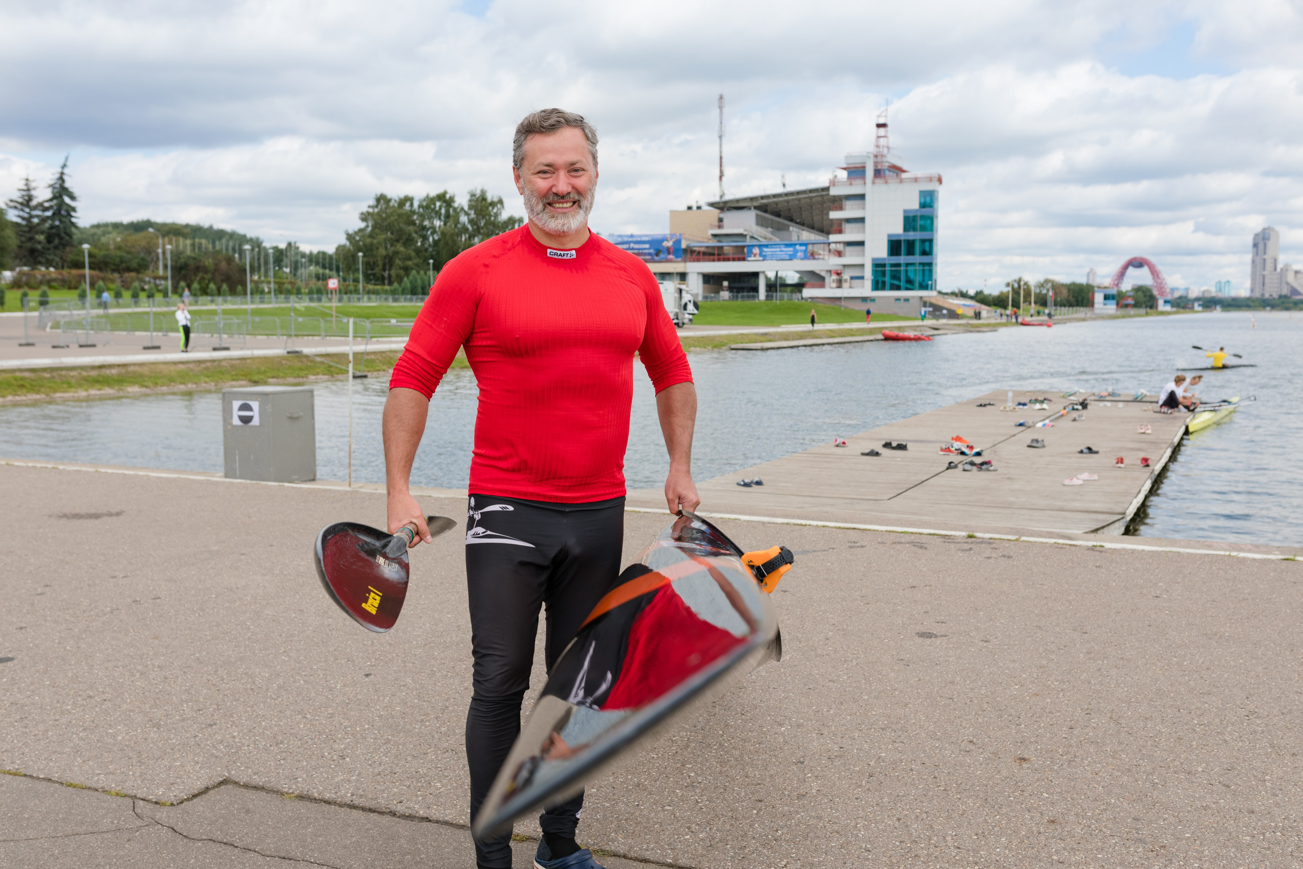 Спортсмен в красной футболке держит лодку в руках на фоне гребного канала