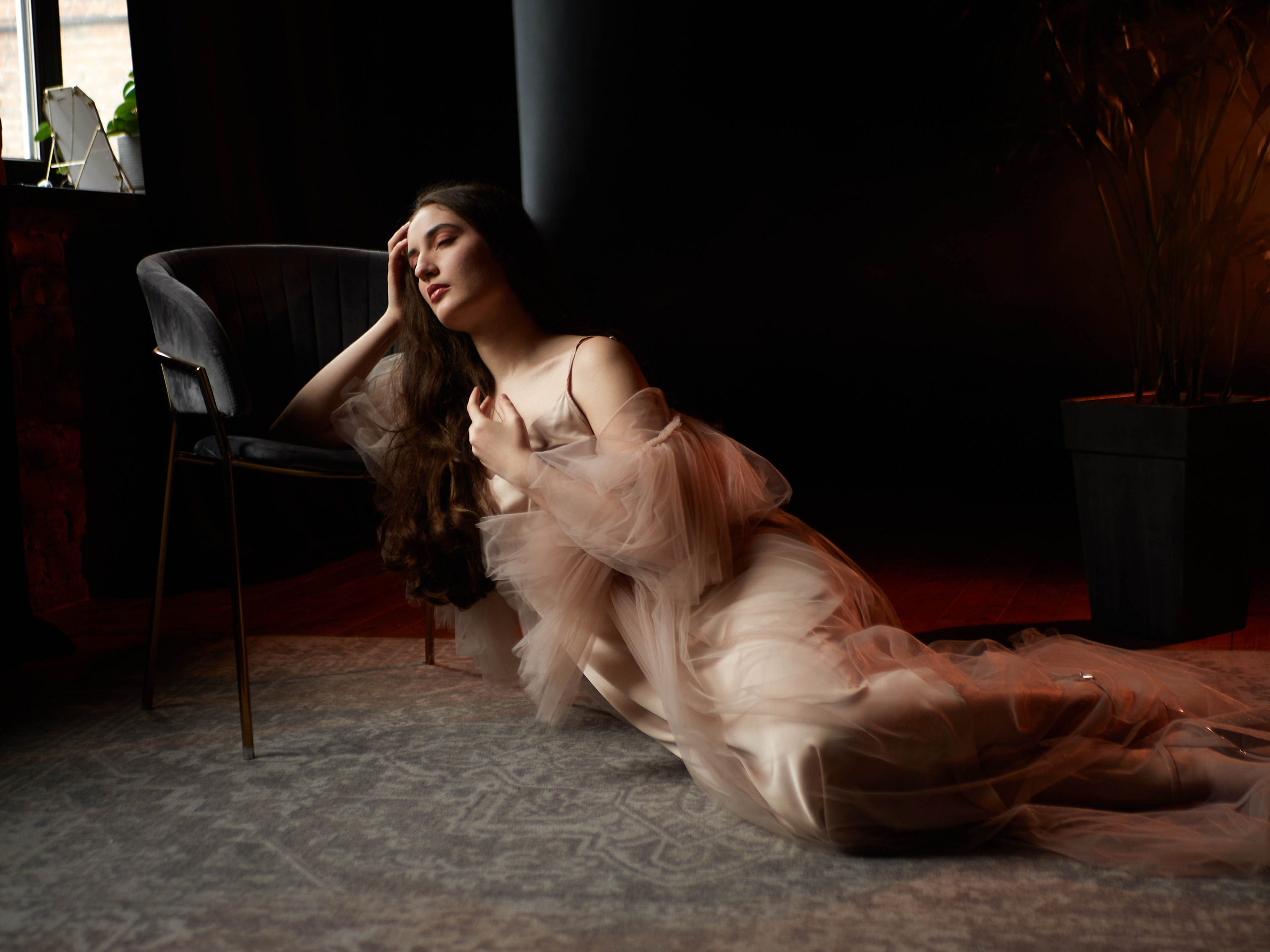 Девушка сидит на полу в бежевом платье на тёмном фоне у окна.