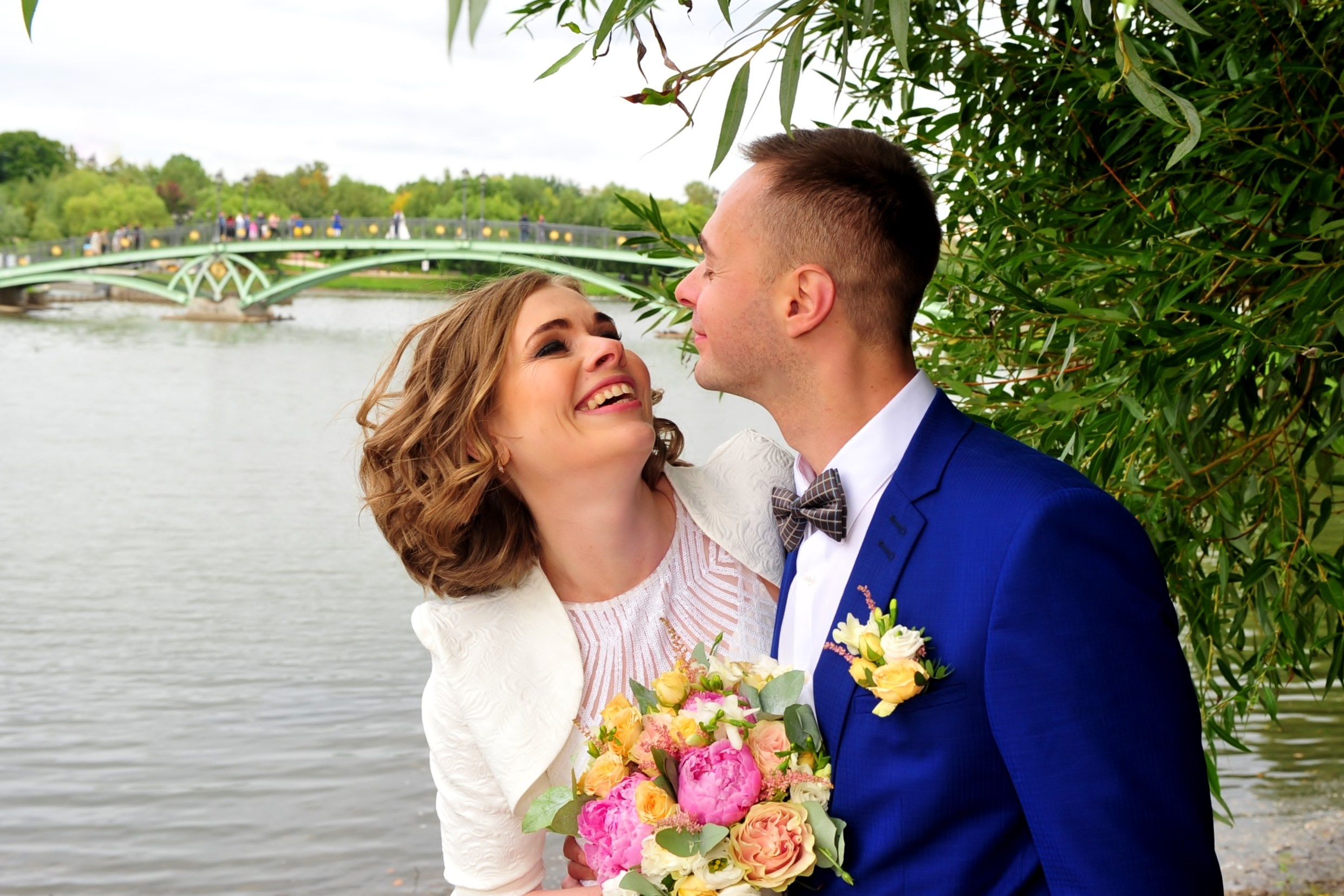 Свадьба в Царицыно хмурым прохладным летним днём, молодожены смотрят друг на друга на фоне пруда, моста и зелени, невеста смеётся