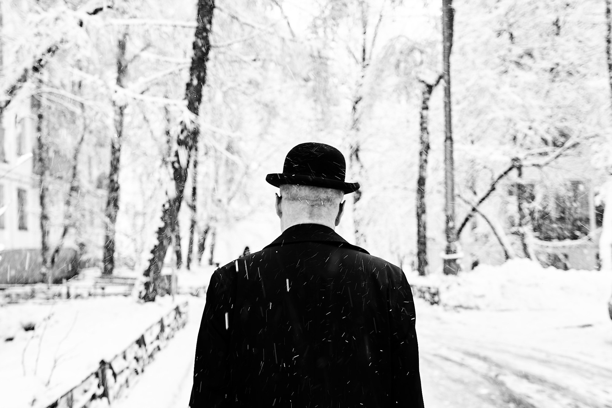 Силуэт мужчины со спины в черном пальто и шляпе-котелке во время снегопада в парке. Черно-белая уличная фотография, ретро-эстетика, зима. Фотограф Виктор Молодцов.