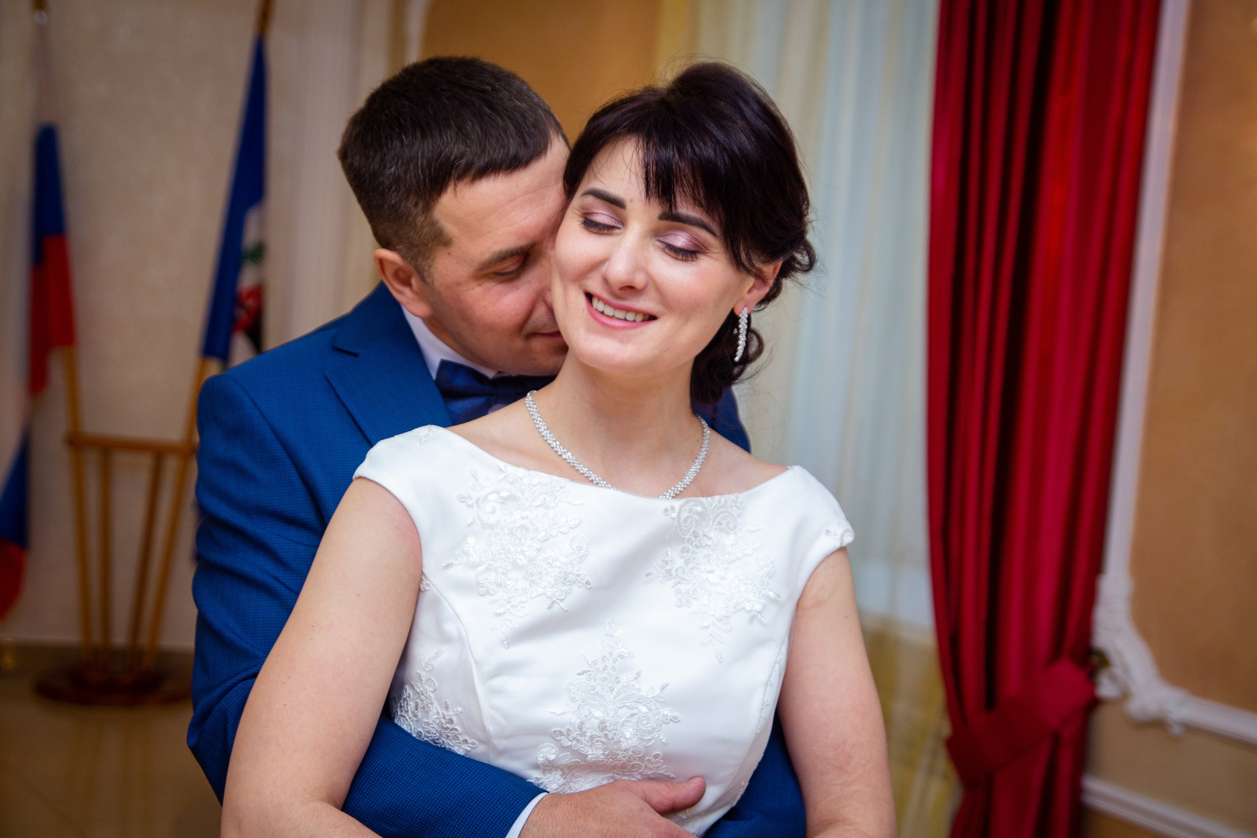 Жених обнимает невесту со спины, оба улыбаются — тёплая эмоция в интерьере ЗАГСа.