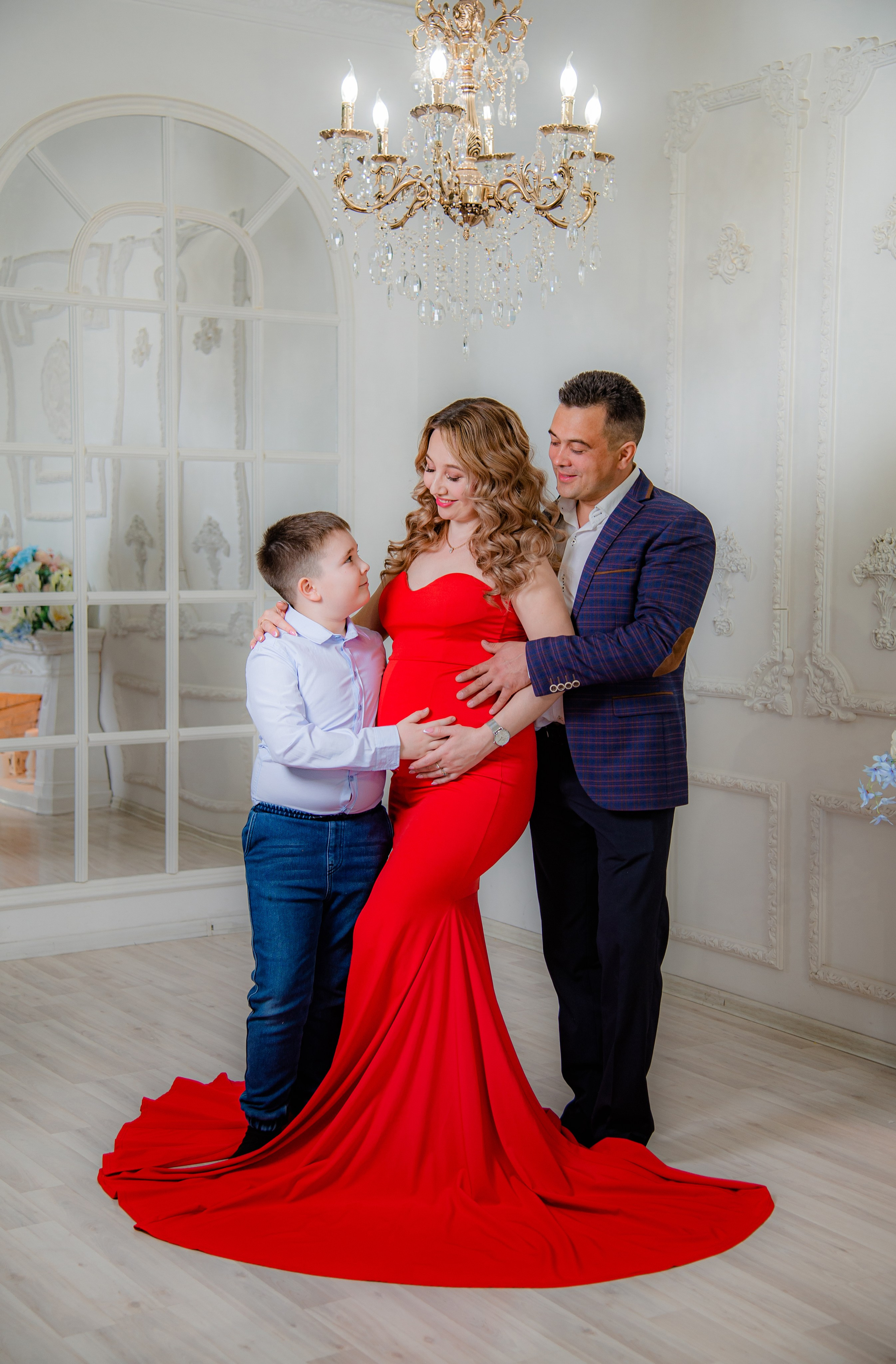 Фотосессия беременных в фотостудии в Уфе: будущая мама в элегантном платье из проката, с профессиональным макияжем и прической. Она счастливо позирует на фоне стильного интерьера, а рядом ее муж и члены семьи. Фотограф помогает в позировании, создавая теплую и уютную атмосферу, отражающую красоту и нежность материнства