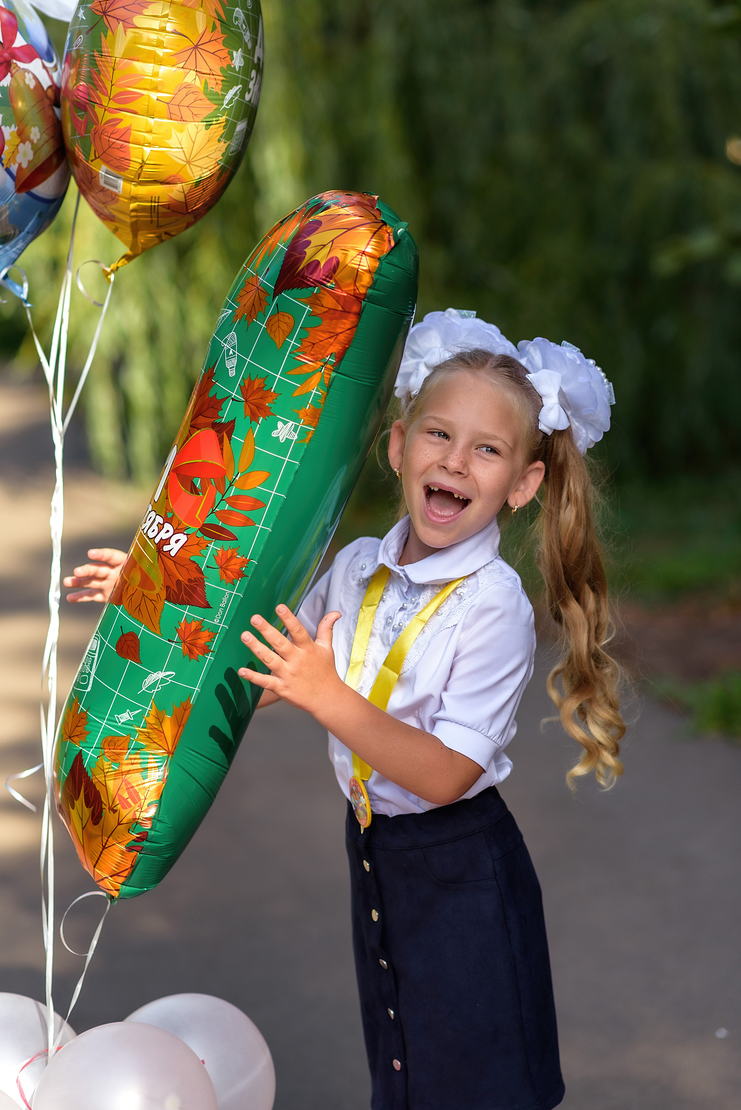 Девочка в школьной форме и с белыми бантами держит шар в форме единицы и улыбается