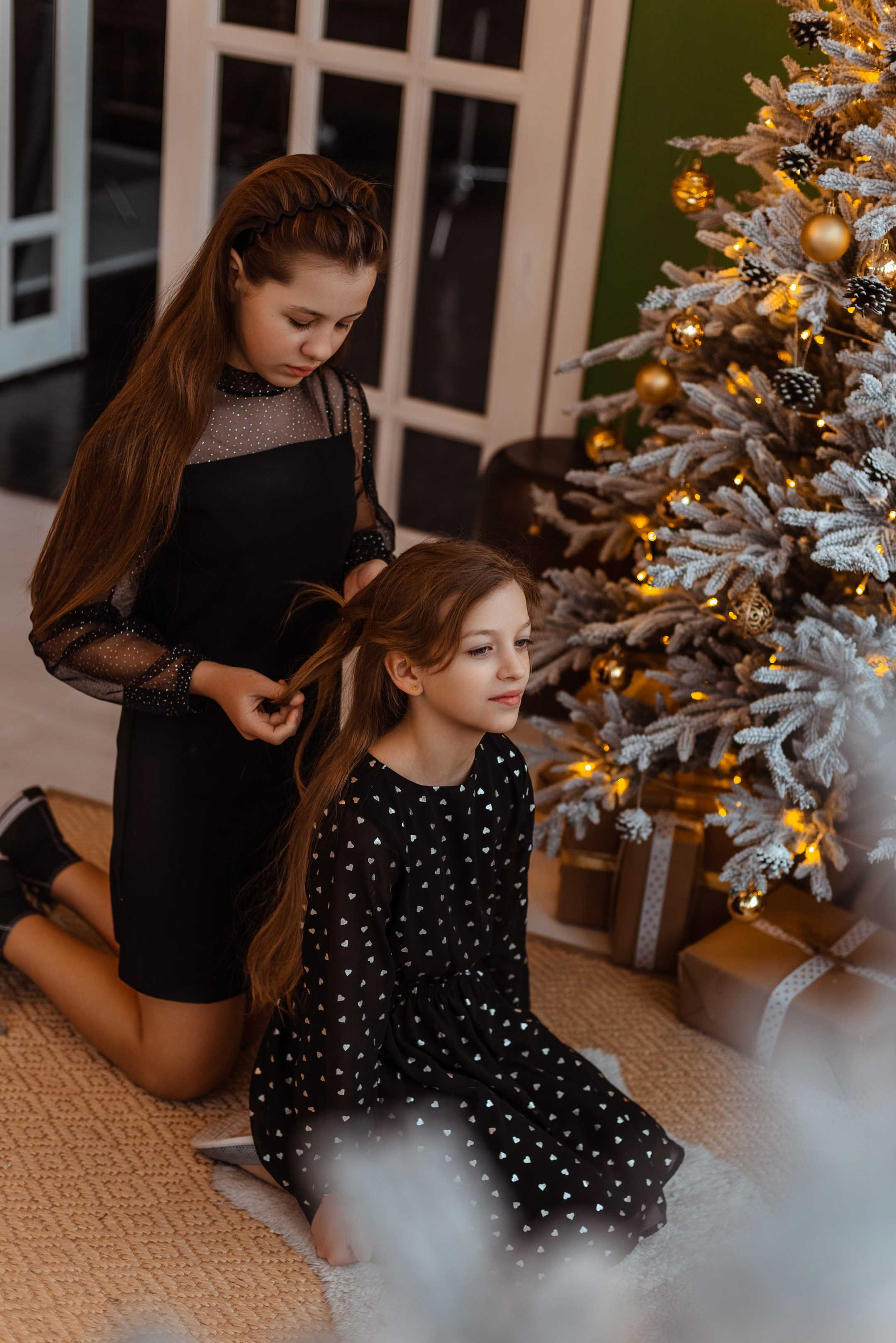 Старшая сестра аккуратно плетёт косичку младшей сестре, создавая атмосферу заботы и любви в преддверии Нового Года