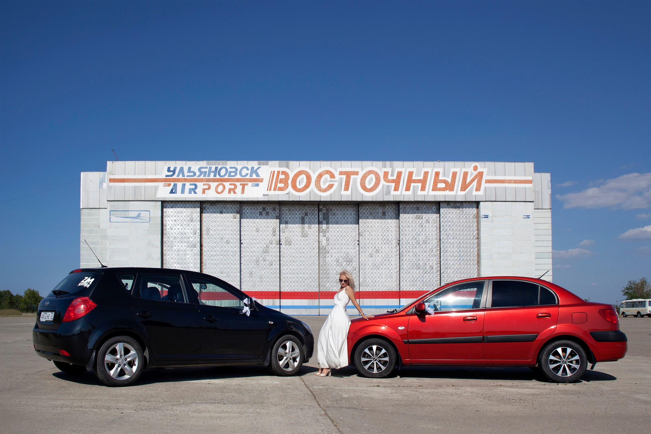 Невеста между красной и черной машинами на фоне здания аэропорта, фотограф Ульяновск