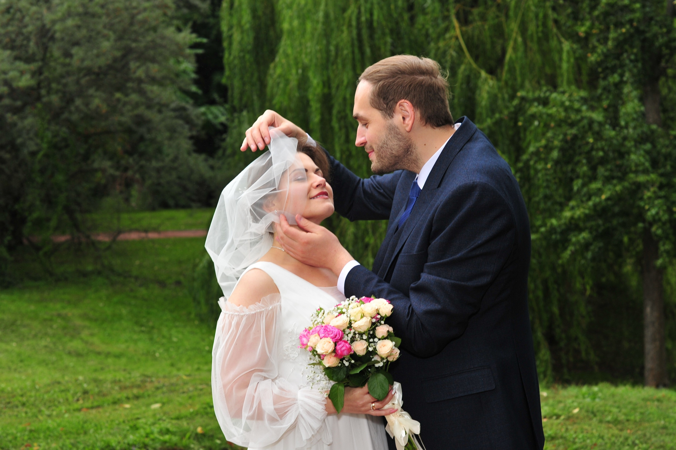 Свадьба в Царицыно пасмурным летним днем, жених приподнимает фату  с лица невесты на фоне плакучей ивы и зелени