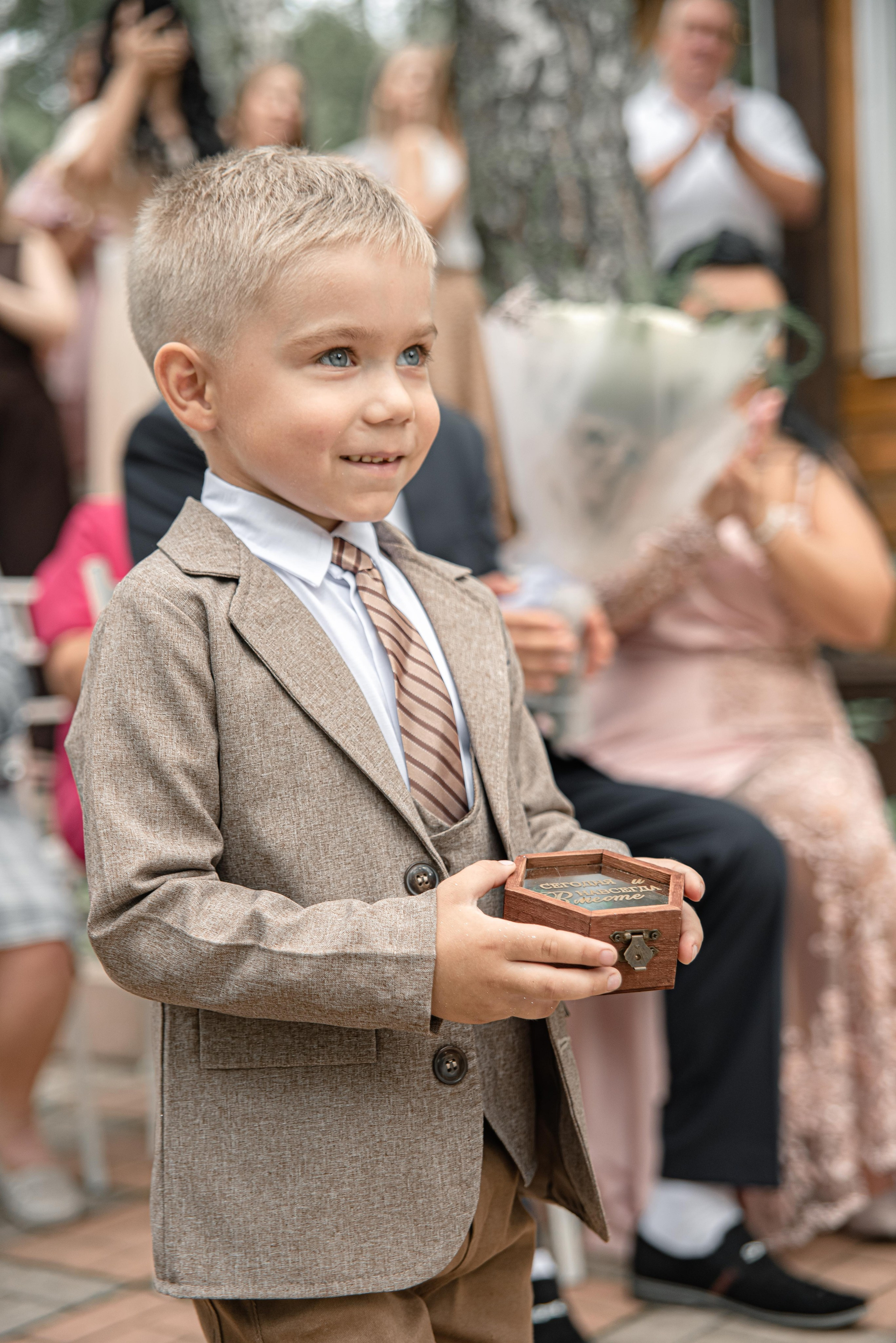 На фото изображен мальчик в элегантном костюме, который держит в руках небольшую деревянную коробочку. Он стоит на фоне гостей, которые наблюдают за происходящим. Атмосфера события наполнена ожиданием и радостью, а вокруг видны элементы свадебного декора и нарядные гости.