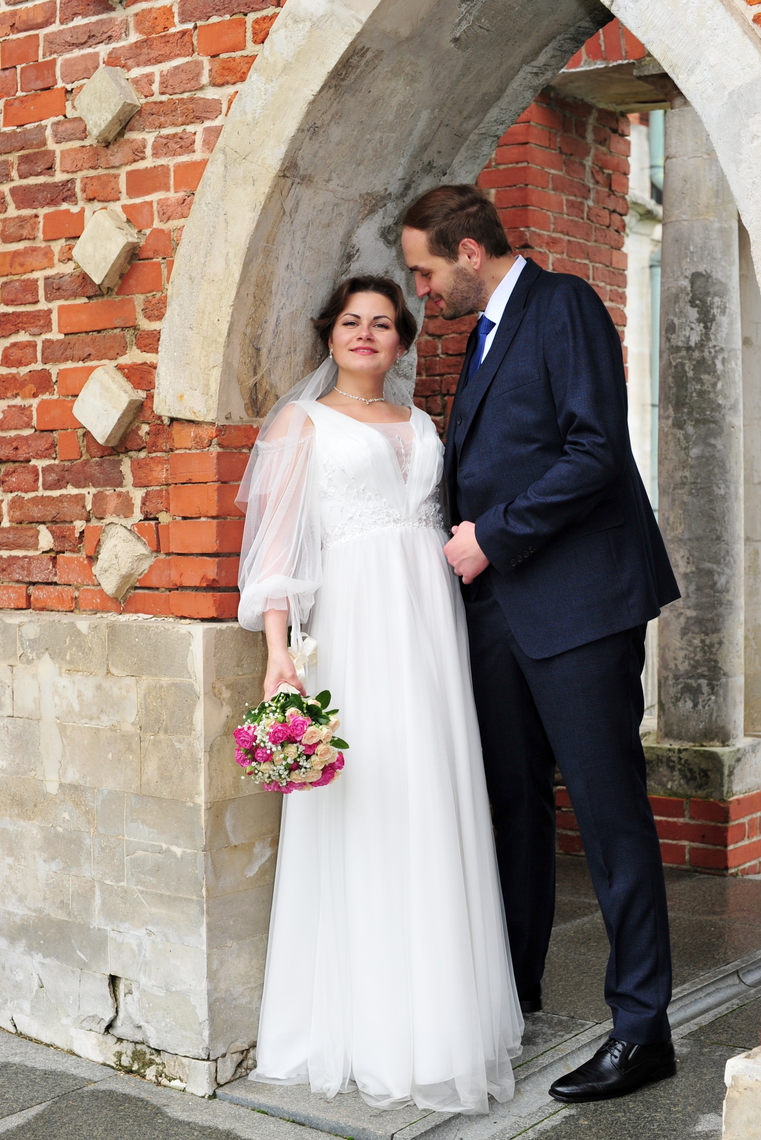 Свадьба в Царицыно пасмурным летним днем, молодожёны стоят рядом и разговаривают в арке из красного кирпича и белого камня 