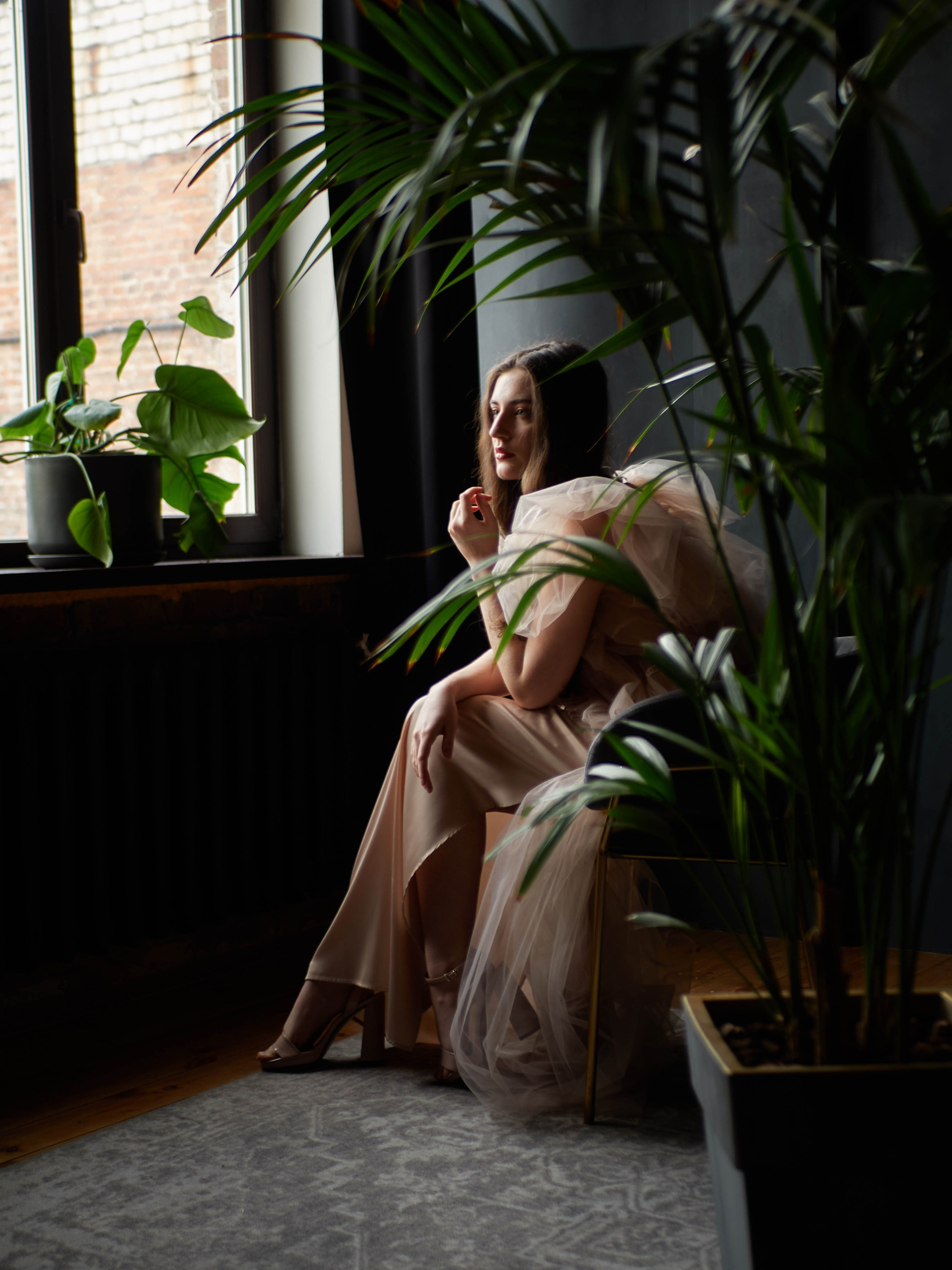 Девушка сидит в кресле в бежевом платье на тёмном фоне у окна.