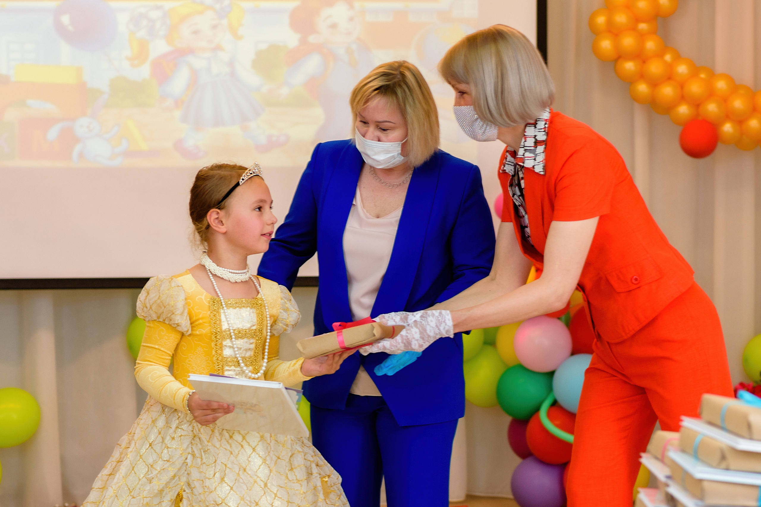 На выпускном в детском саду девочке вручают грамоту заведующая и воспитатель