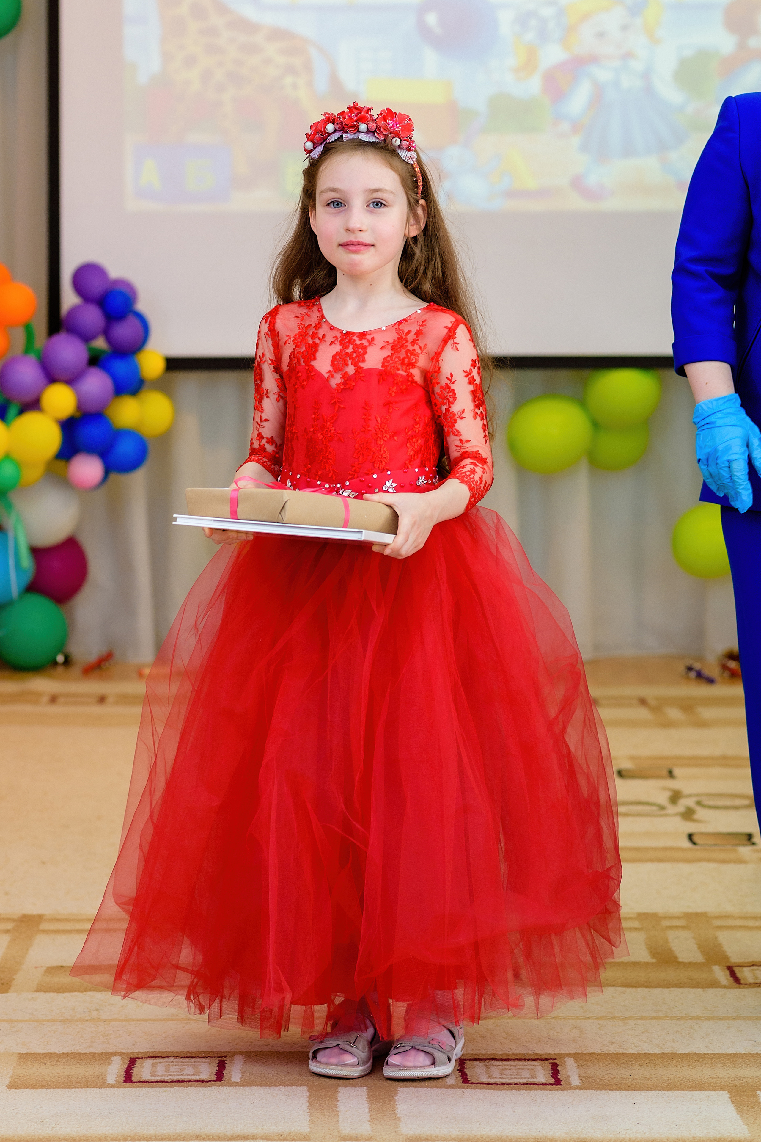 Девочка в красном платье стоит в музыкальном зале и держит подарки