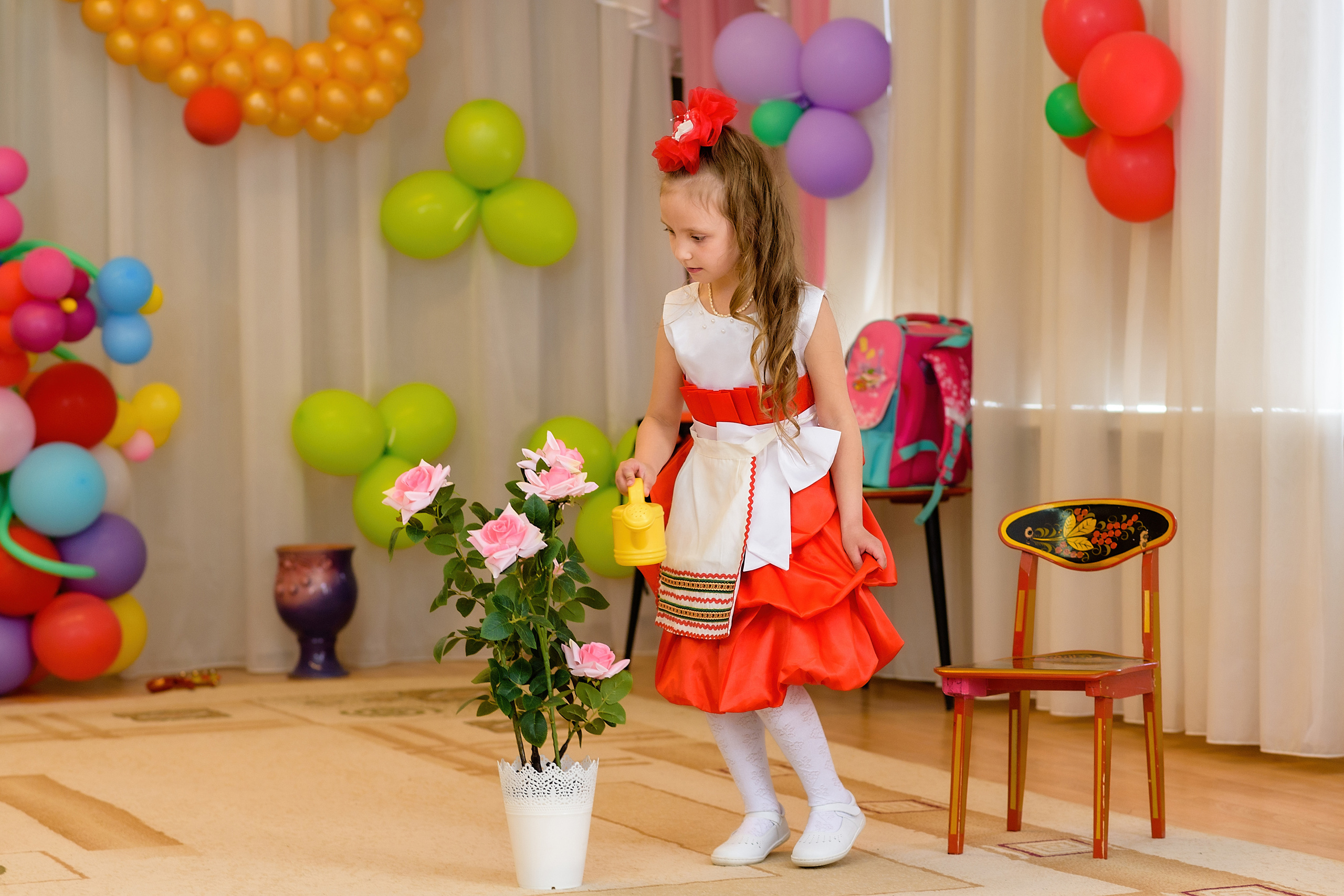 Девочка в красном платье участвует в сценке на выпускном из детского сада. Она поливает цветы