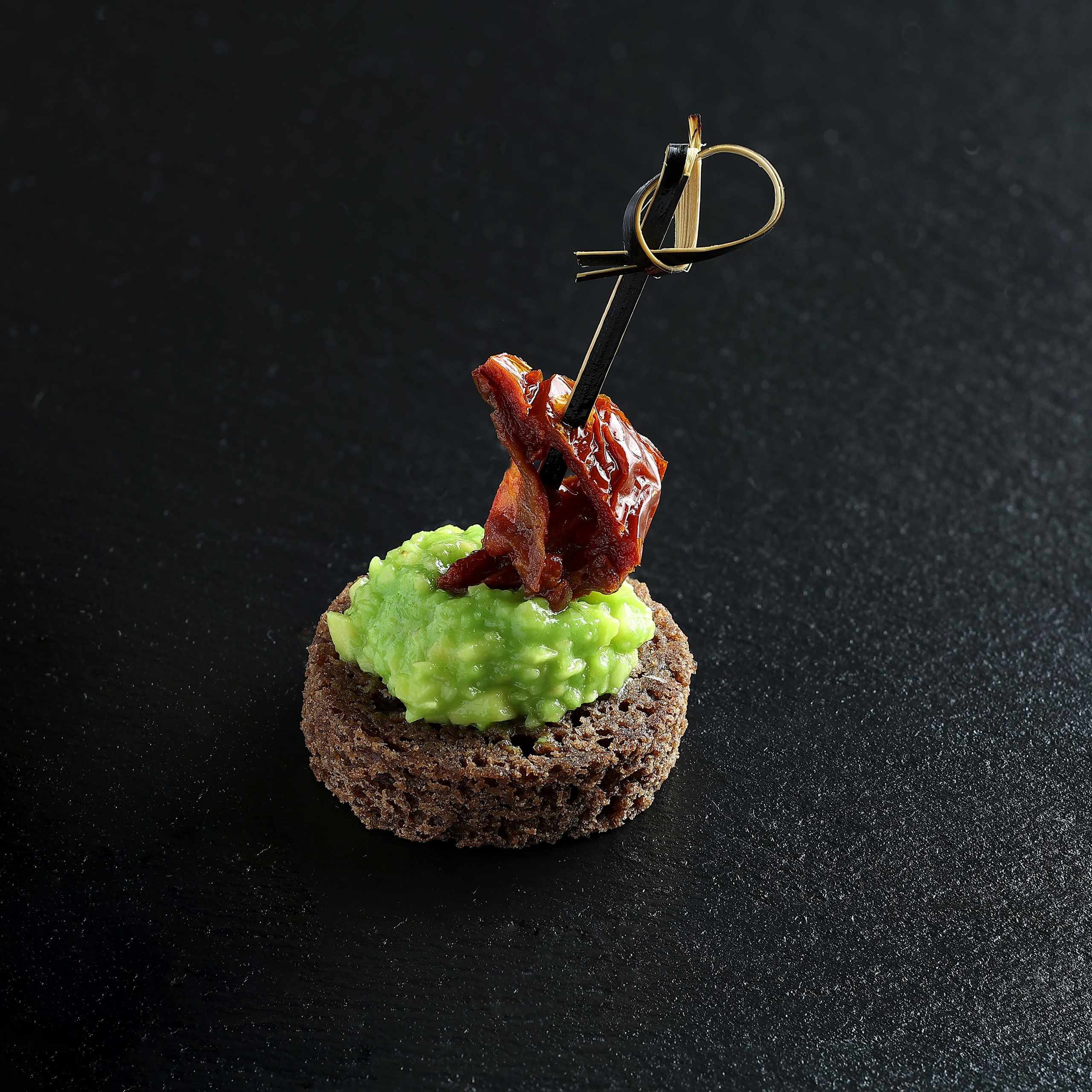 Закуска на бородинском хлебце с гуакамоле и вялеными томатами, проколотая шпажкой, на тёмном фоне.