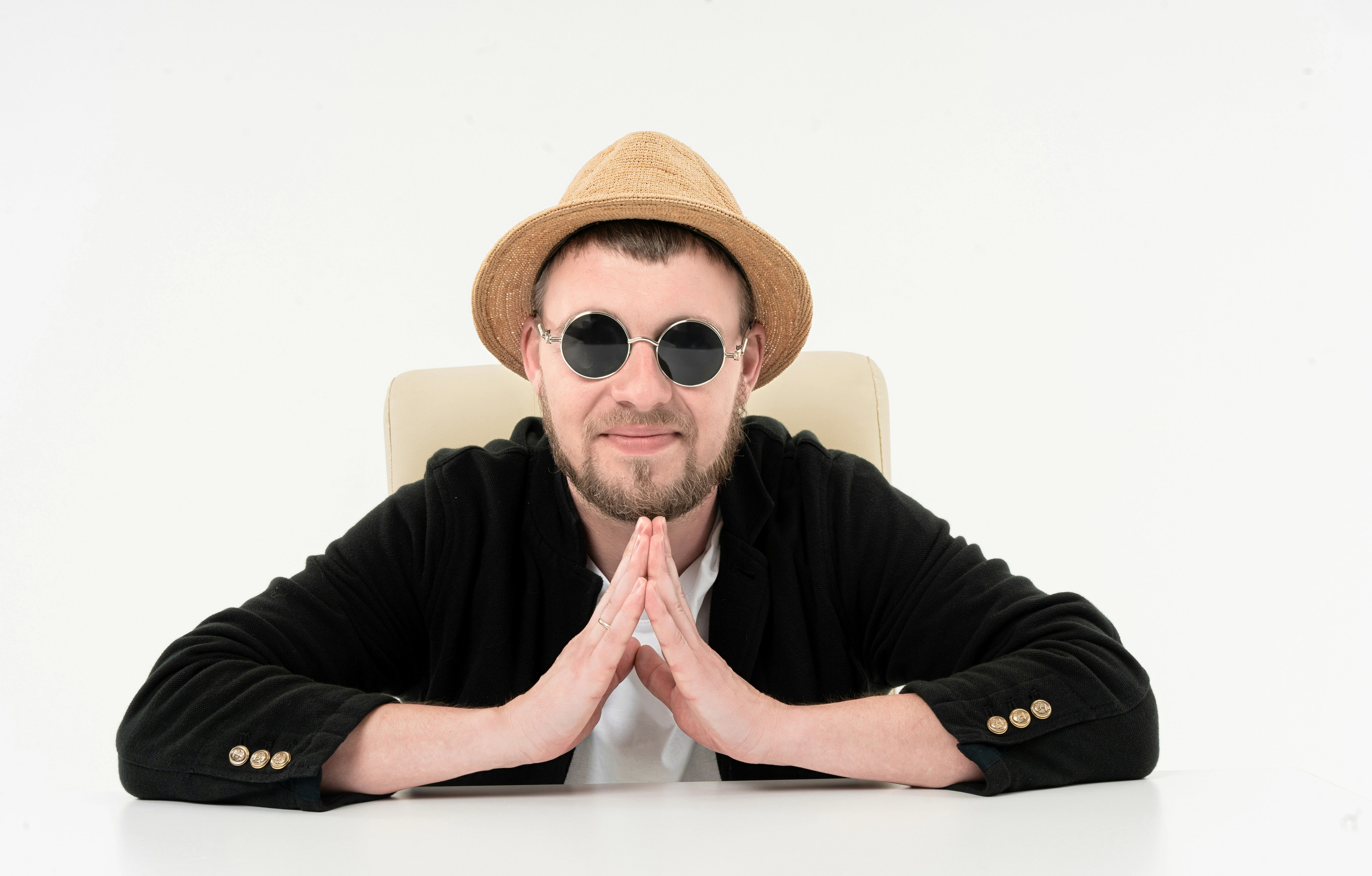 На данной фотографии изображен человек, сидящий за столом с скрещенными руками в жесте, который может указывать на размышление или умольение. Он одет в соломенную шляпу, черный свитер с пуговицами на манжетах и белую футболку. Человек находится перед однотонным фоном.