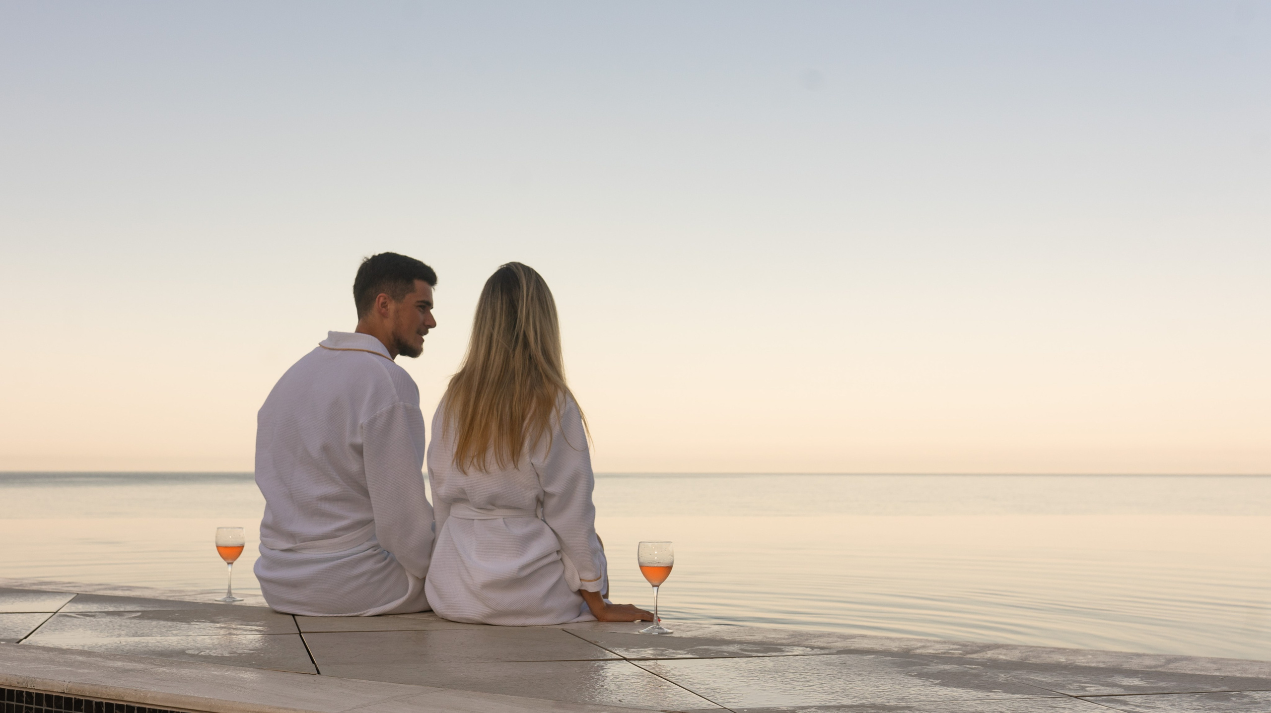 Мужчина и женщина сидят на краю пейзажного бассейна, глядя на океан. Мужчина и женщина оба одеты в белые халаты, а волосы женщины собраны в конский хвост. Мужчина держит бокал красного вина в левой руке и смотрит на женщину. Женщина держит бокал красного вина в правой руке и смотрит на океан. На заднем плане садится солнце.