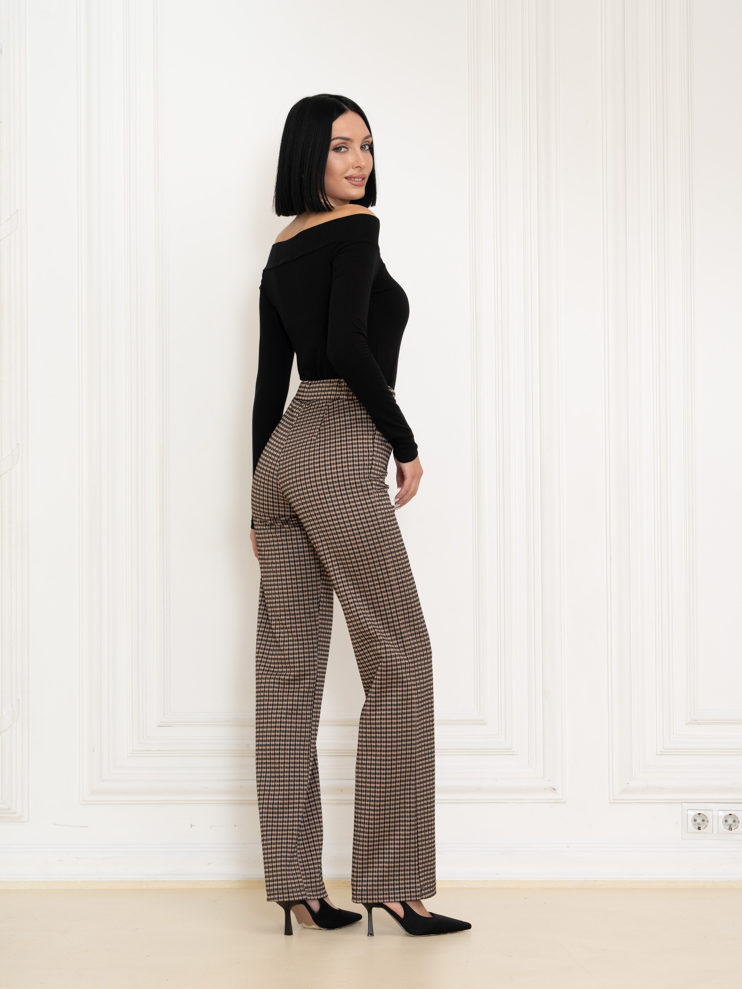 Девушка брюнетка стоит вполоборота в черном боди и брюках в интерьере