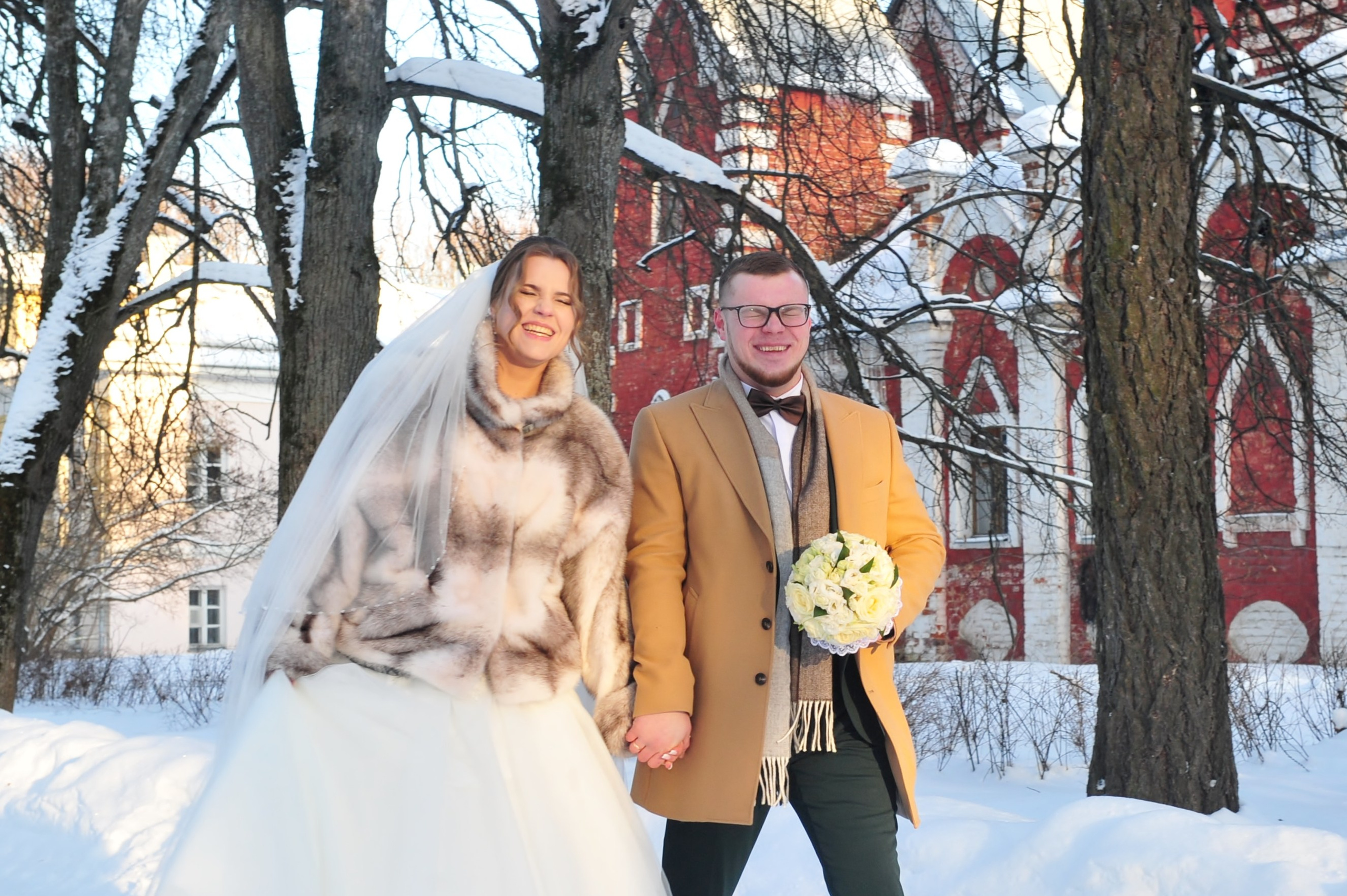 Зимняя свадьба в усадьбе Суханово в сильный мороз, жених и невеста идут по  заснеженной дороге и держатся за руки на фоне красно-белой усадьбы