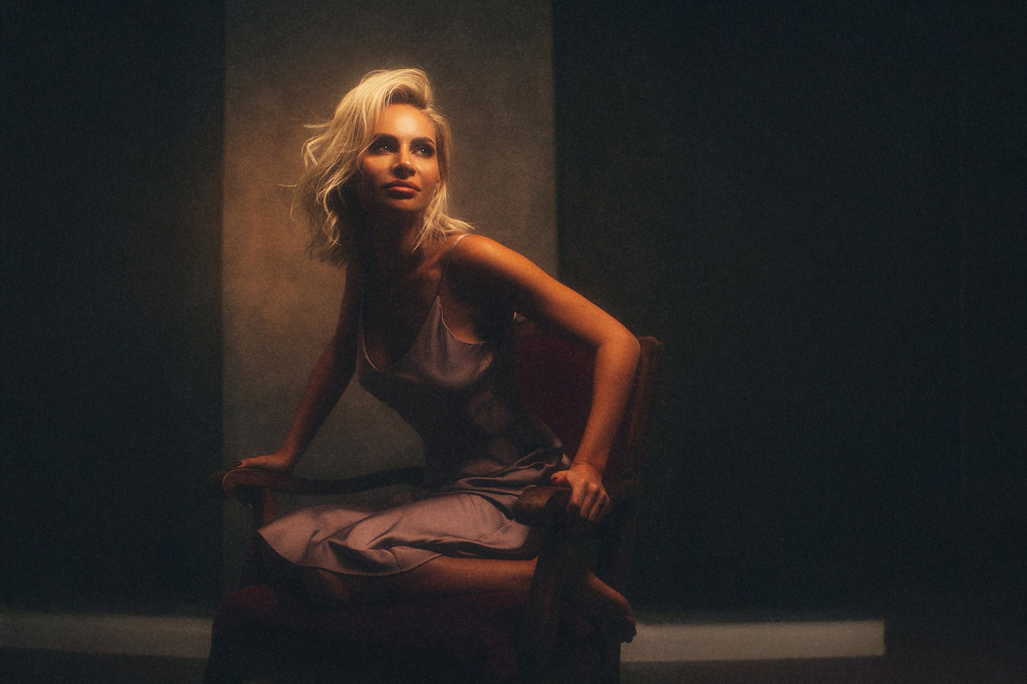 Атмосферный портрет блондинки в шелковом платье, сидящей в винтажном кресле. Теплый, мягкий свет и легкий эффект дымки создают кинематографичный и чувственный образ. Студийный фотограф Галина Скорик