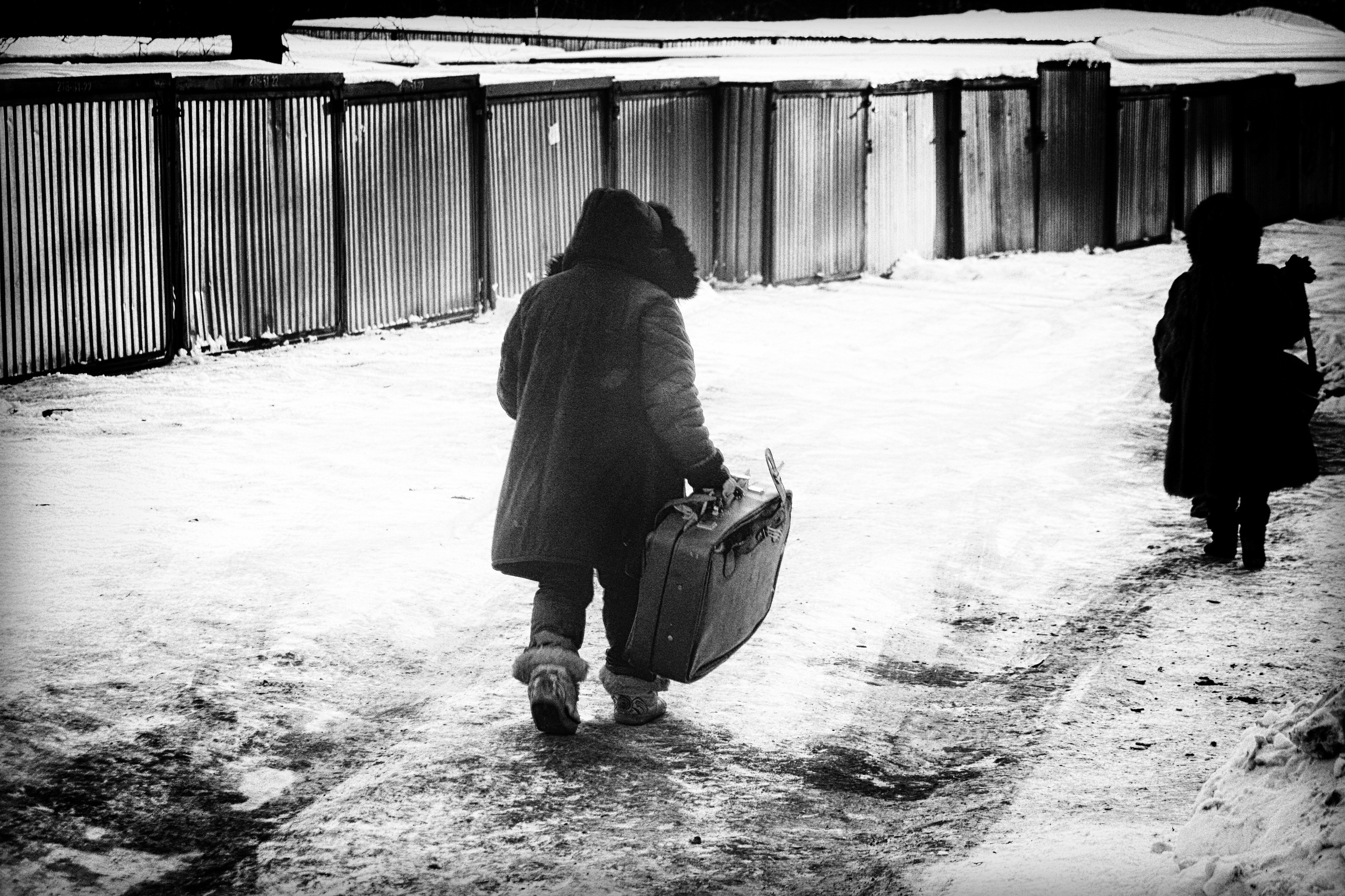 Человек с чемоданом идет по заснеженной дороге мимо гаражей, зимний городской пейзаж, черно-белая уличная фотография о пути