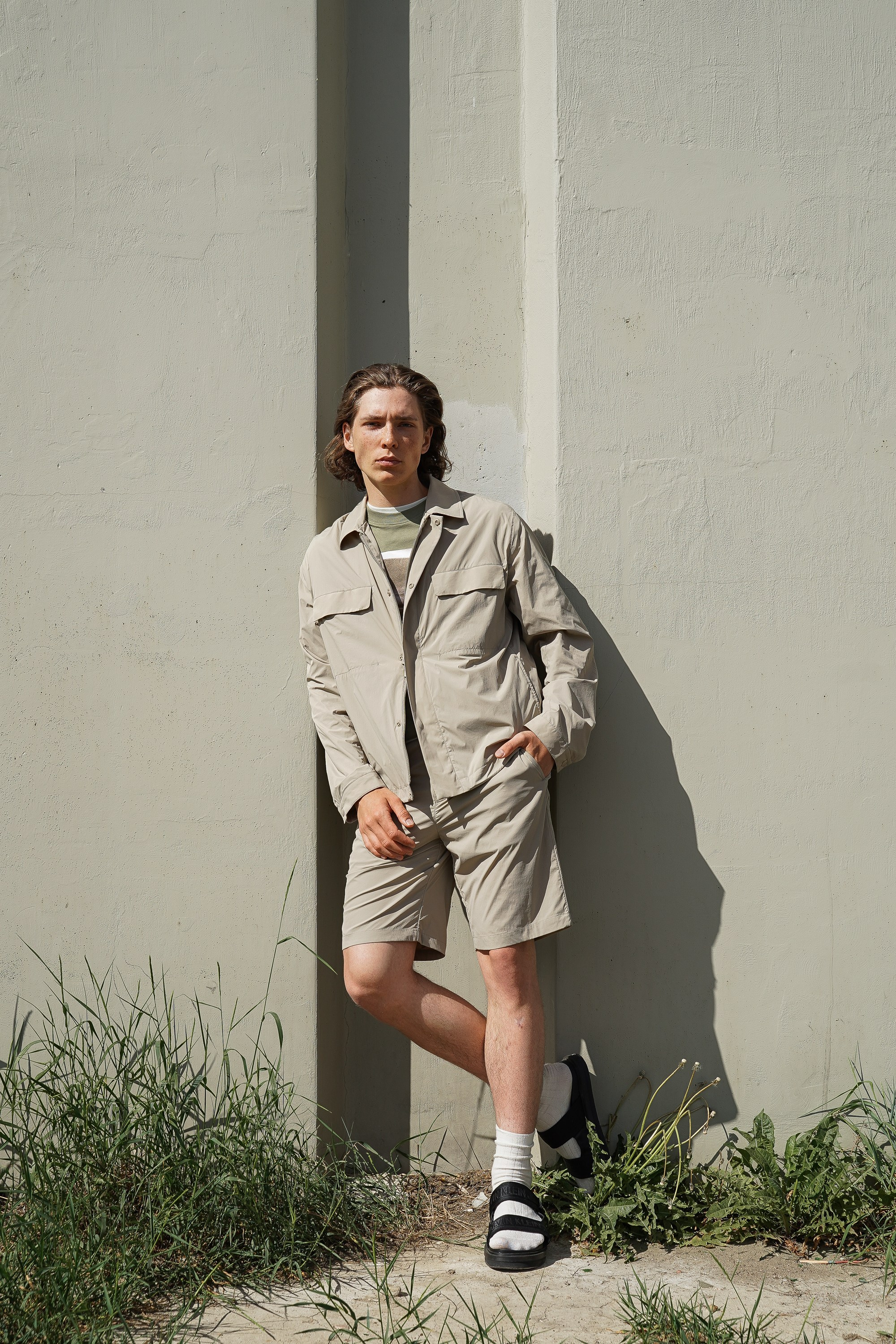 Парень модель с длинными волосами в куртке и шортах стоит у стены