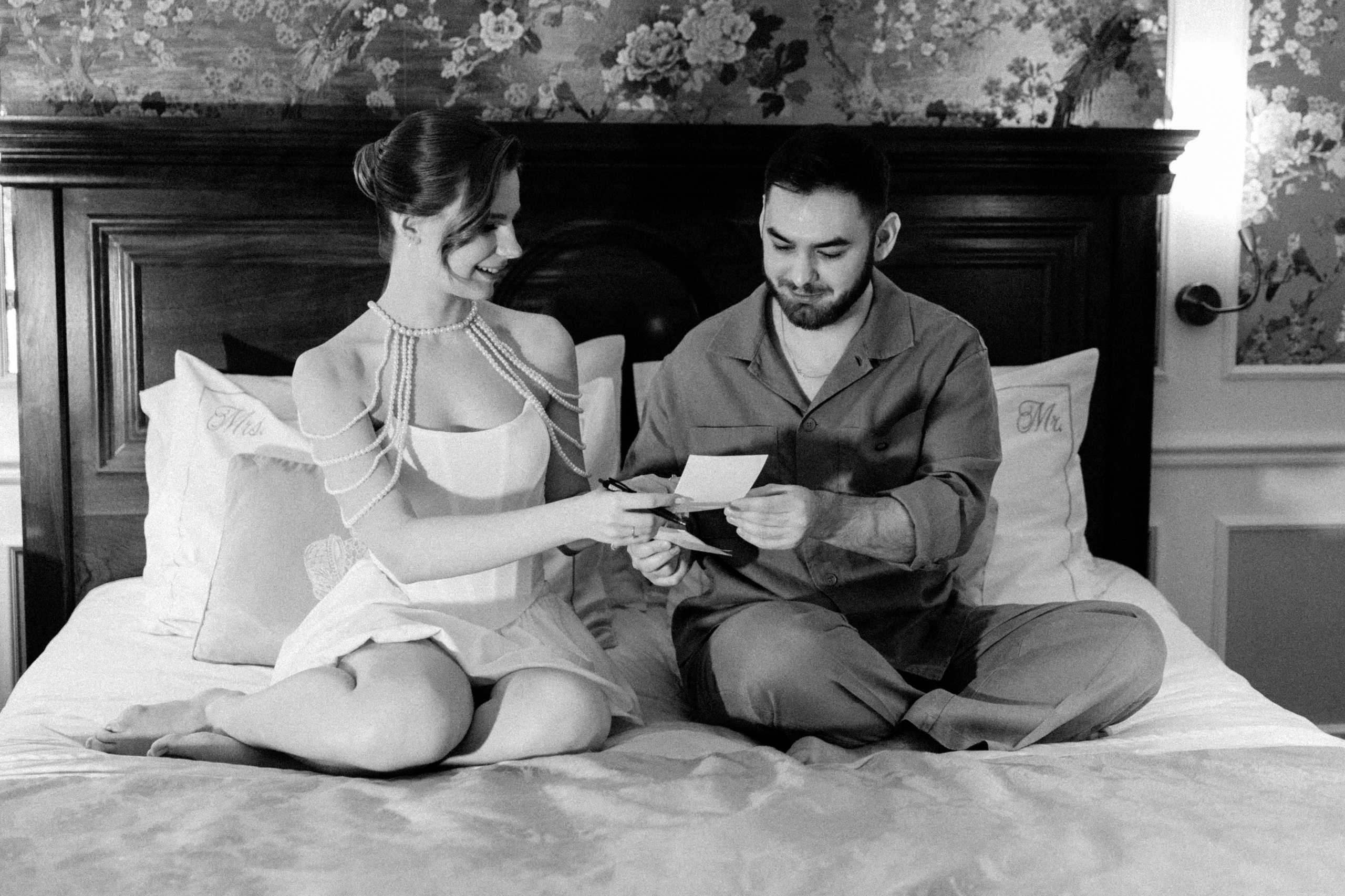 Черно-белое фото жениха и невесты, сидящих на кровати и обнимающихся письмами. 