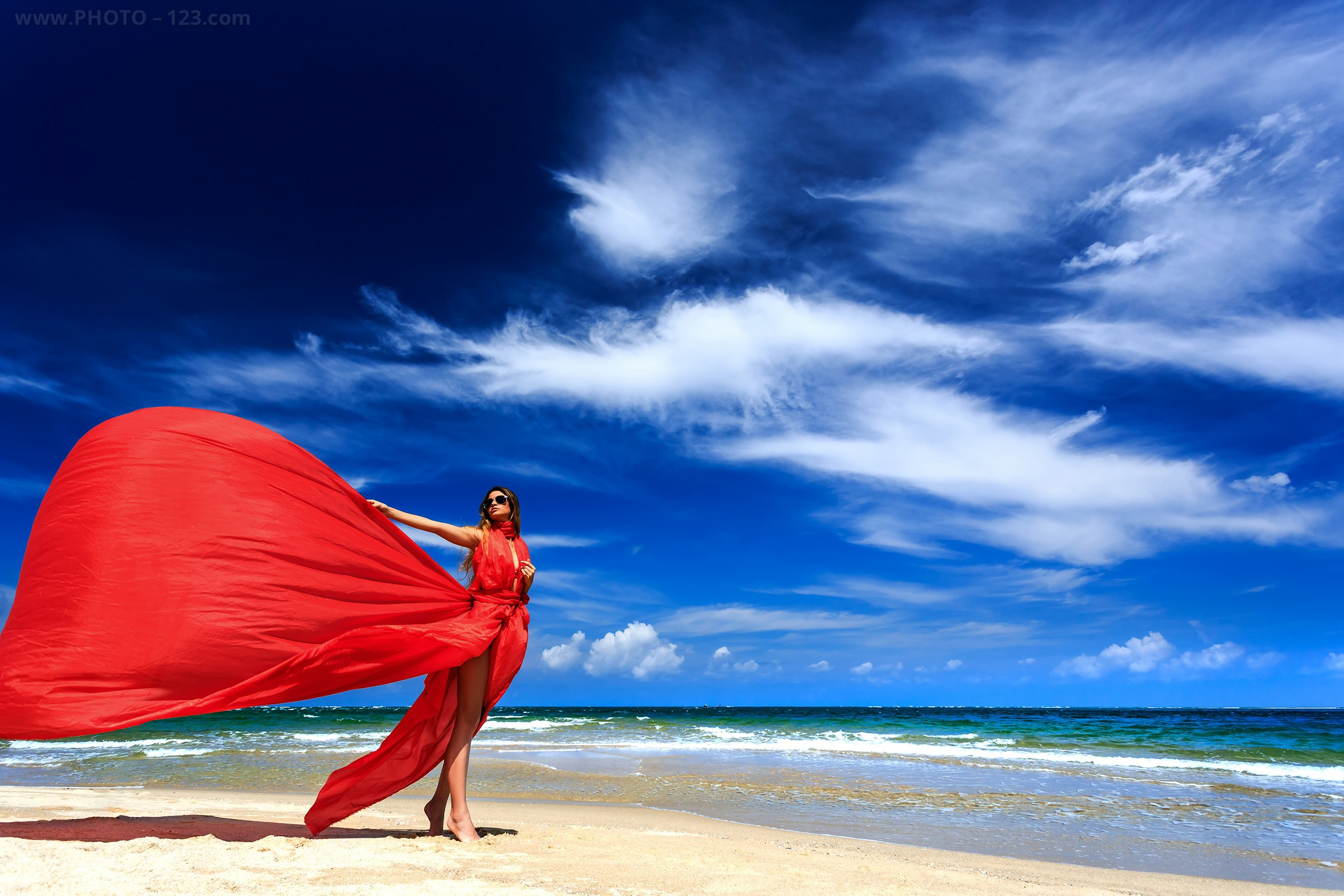 Fashion portrait of a woman in a flowing red dress standing on a beach under a deep blue sky, photographed in natural light in Phu Quoc