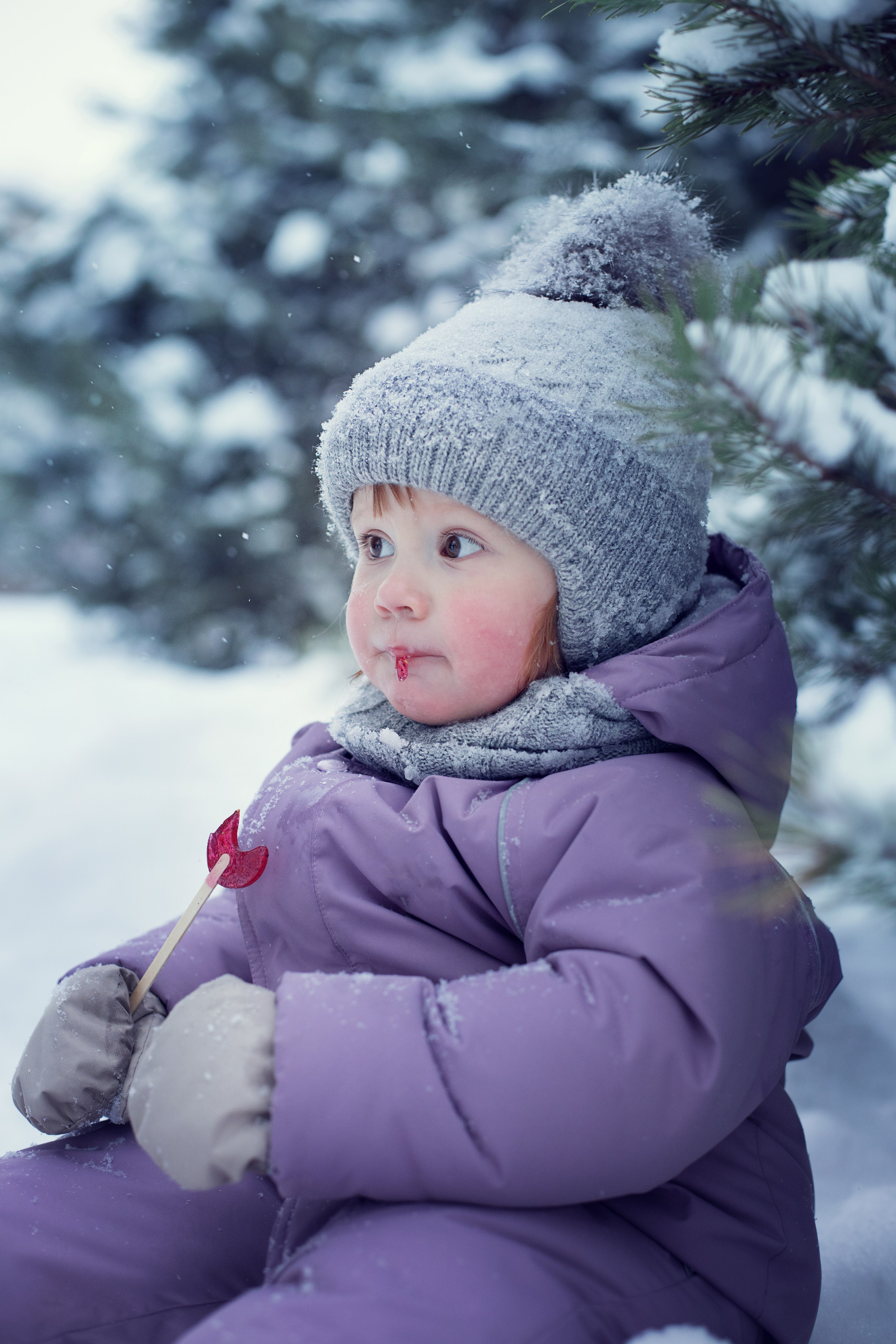 Девочка в лиловом комбинезоне сидит с зимнем лесу и ест леденец