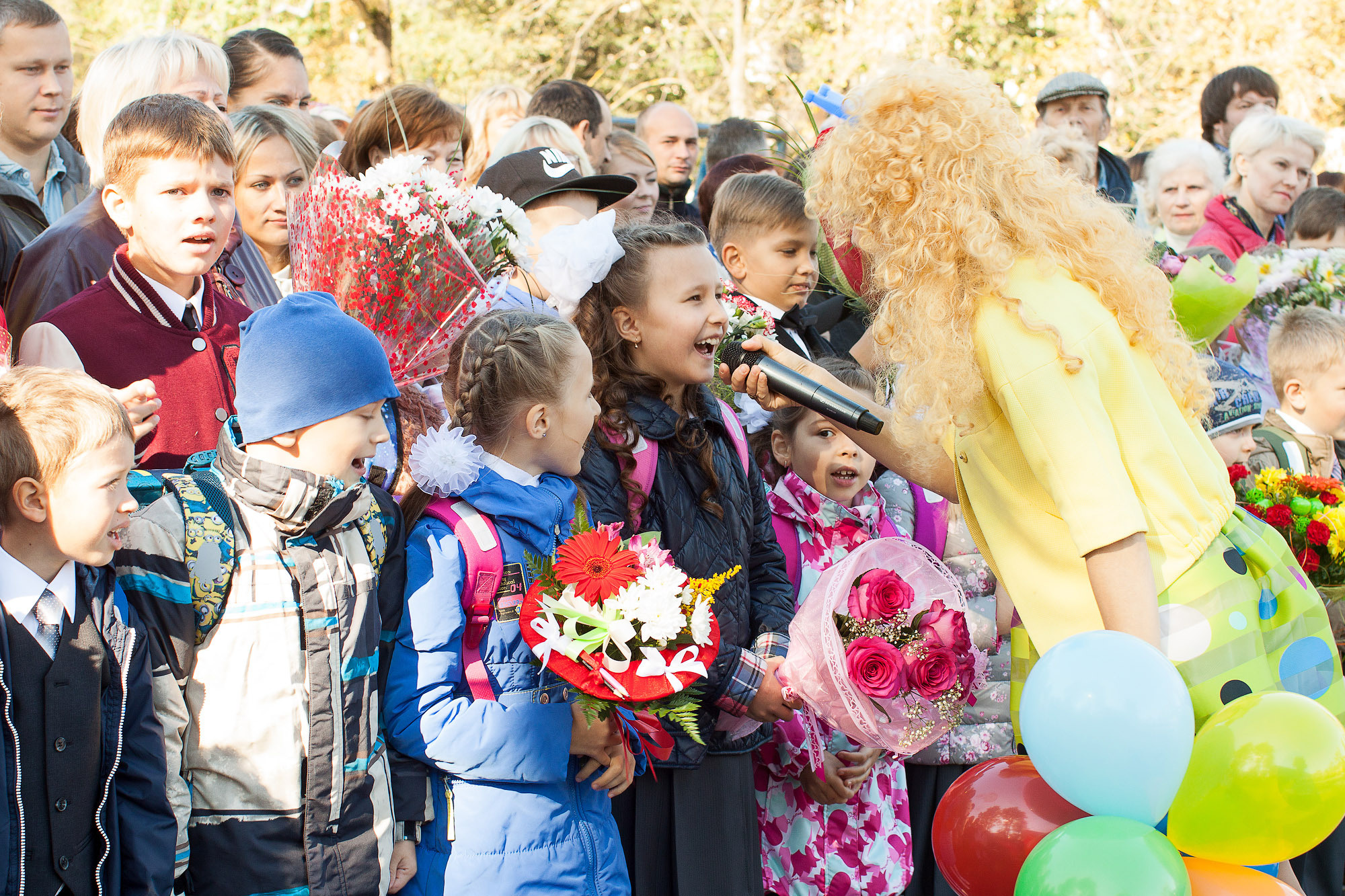 Аниматор в желтом костюме на линейке 1 сентября берет интервью у первоклассника.
