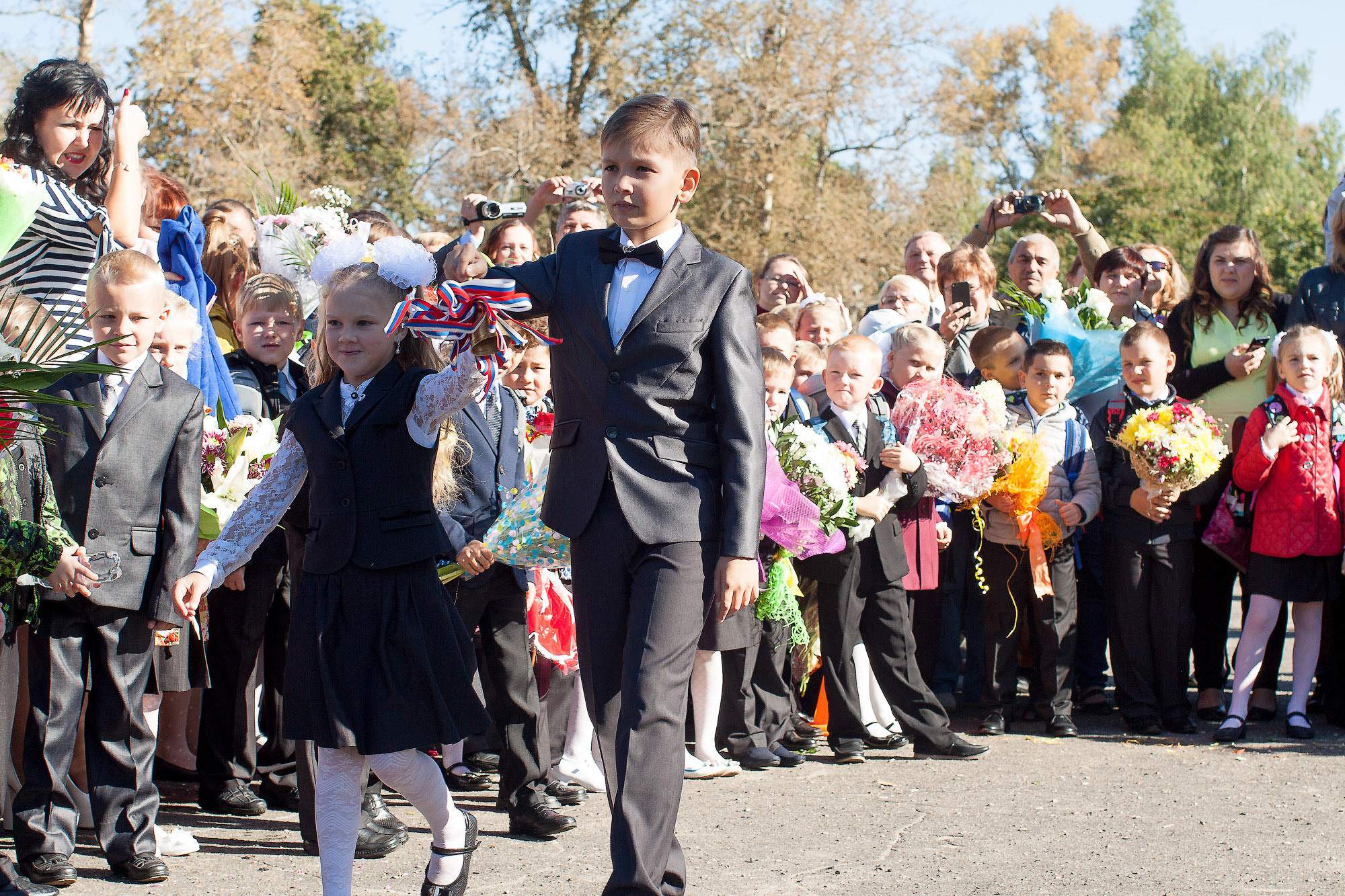Мальчик 4 класса и девочка 1 класса подают последний звонок на линейке на улице.