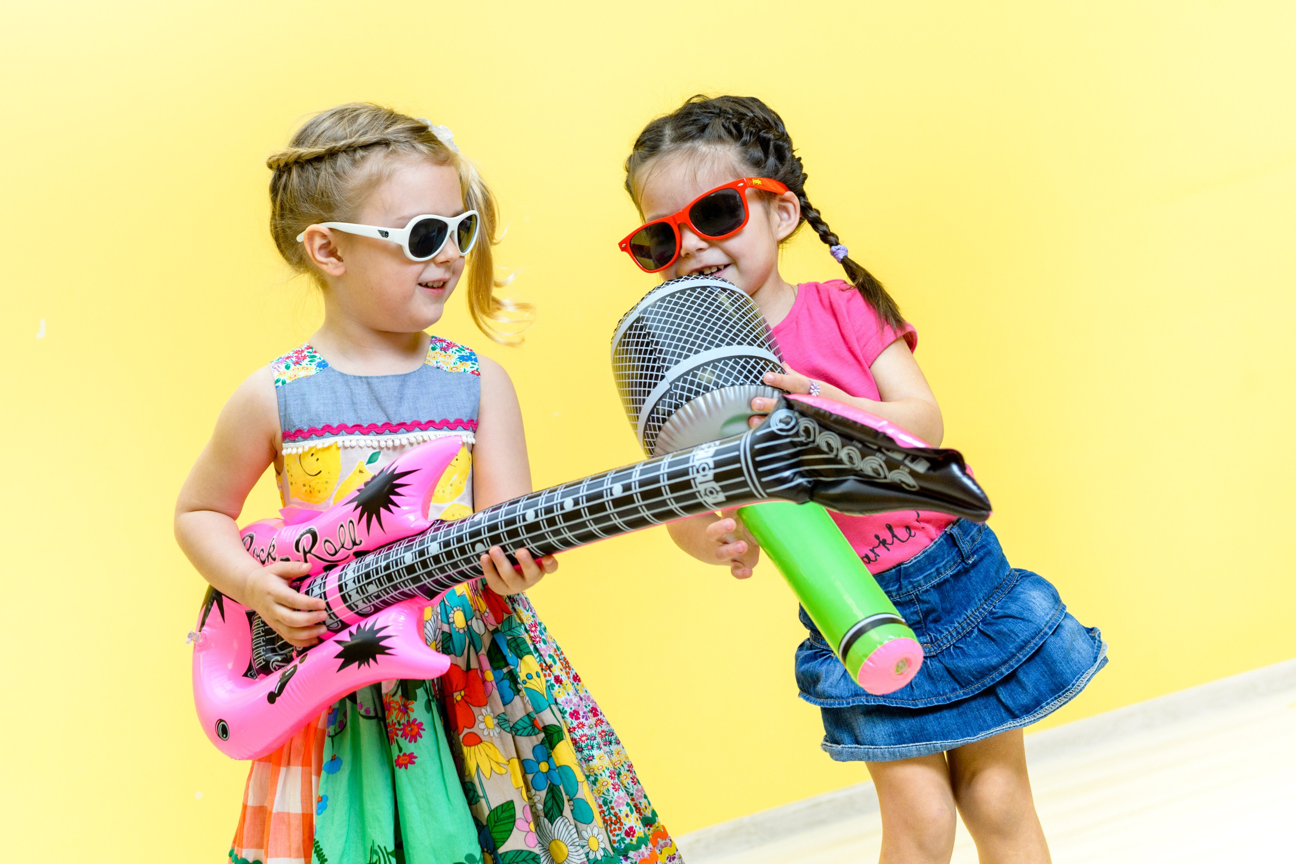 Две девочки в солнцезащитных очках позируют на ярком жёлтом фоне в ярких платьях с надувными микрофоном и гитарой.