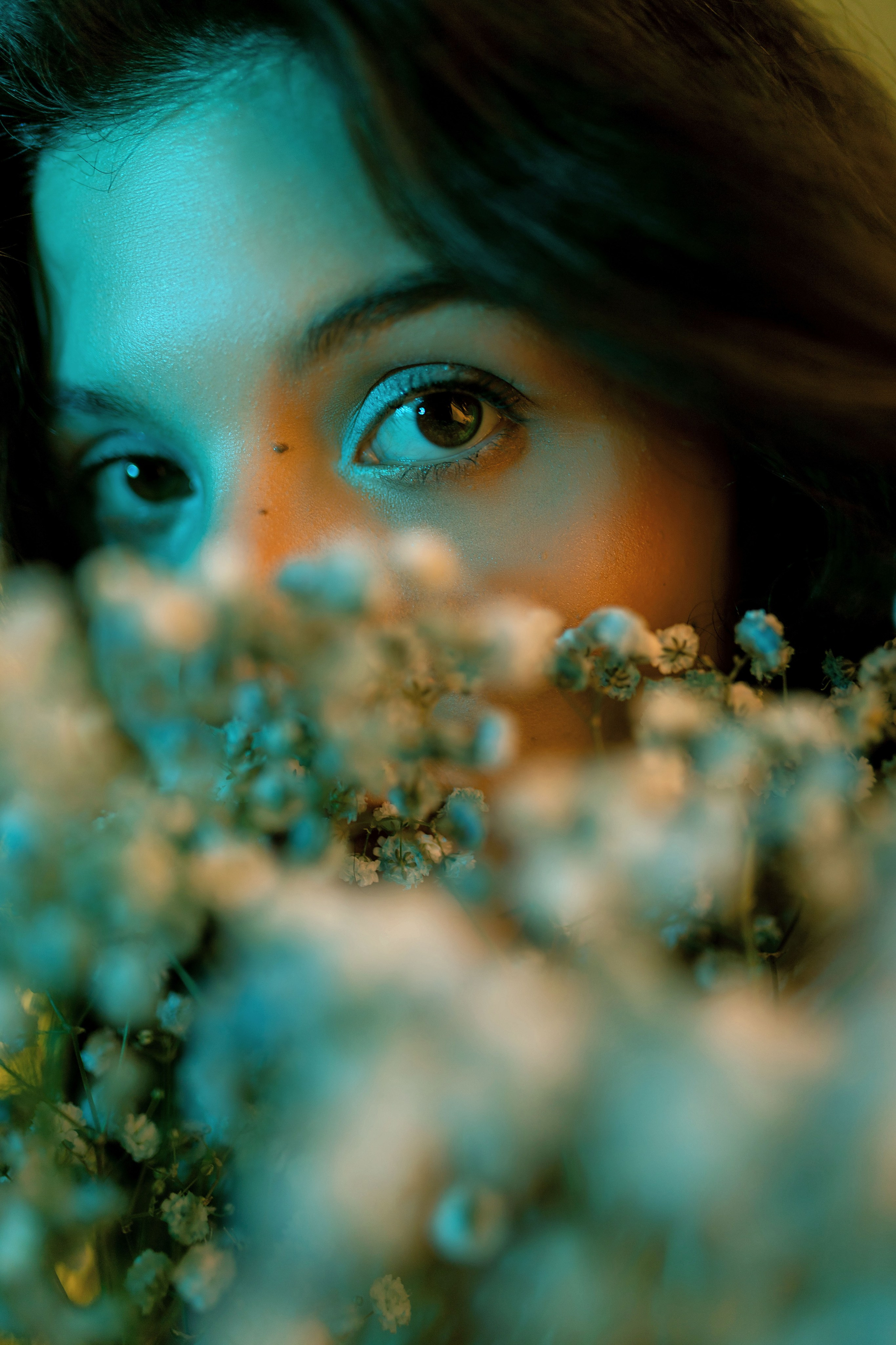 Молодая женщина частично скрыта букетом нежных цветов, но видны ее выразительные глаза, освещенные мягким и успокаивающим светом, создающим чарующую атмосферу.