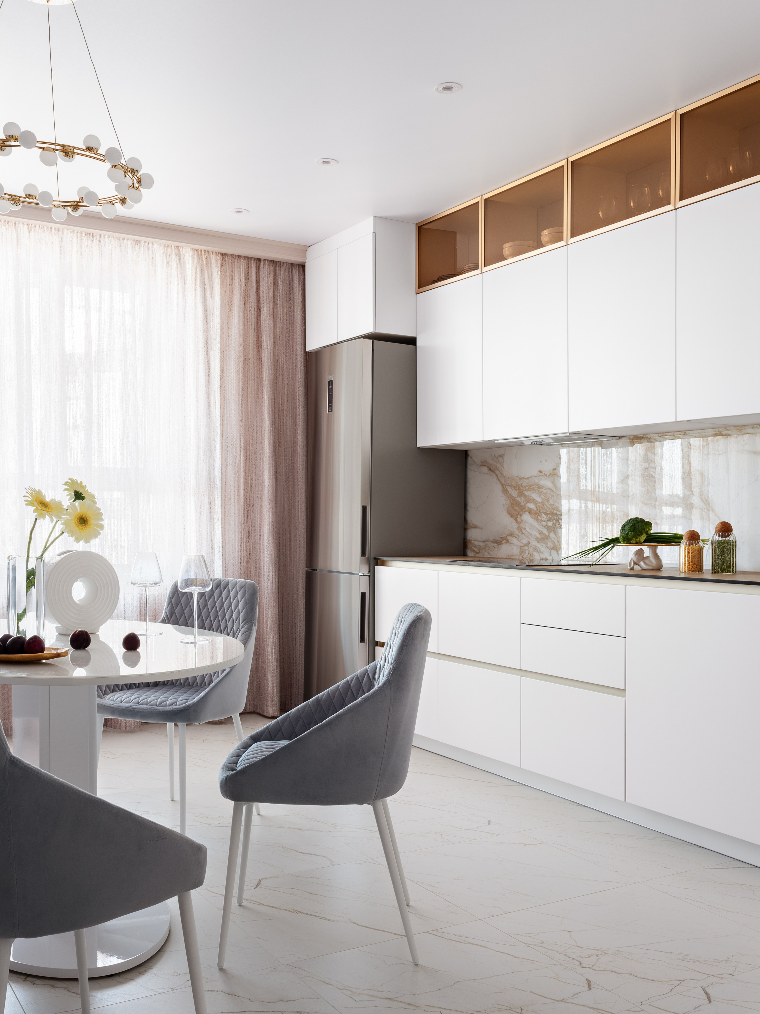 Общий вид кухни-столовой: белые фасады кухни, мраморный фартук, современная люстра и уютная обеденная зона с мягкими стульями и круглым столом; интерьер разработан дизайнером Натальей Бунеевой, стилист Наталья Якушева, фотограф Сергей Денисенко.