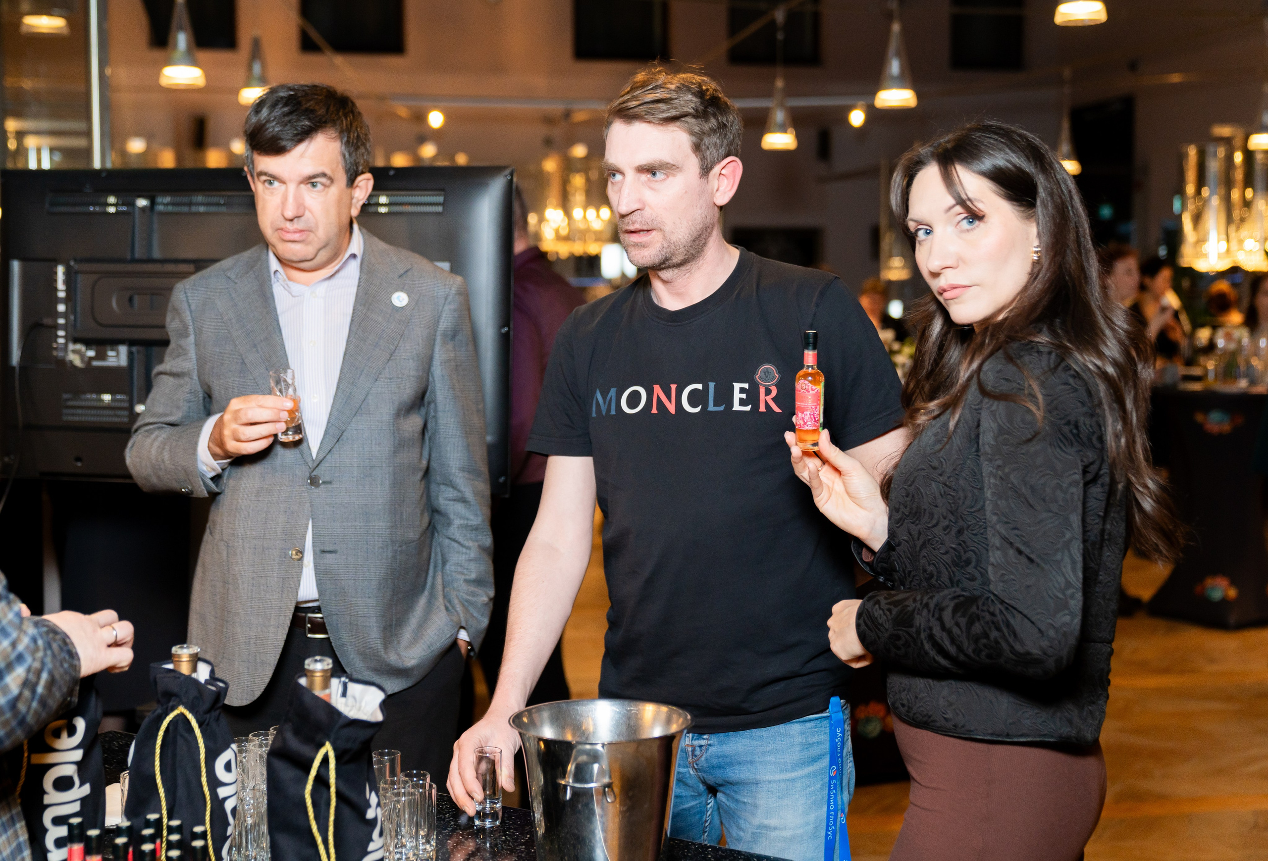 Фотография с вечеринки в отеле Radisson Sonya. Мужчина в сером пиджаке, мужчина в чёрной футболке Moncler и женщина с мини-бутылочкой ликёра у барной стойки. Автор: Евгений Мальцев (evrednix.ru).