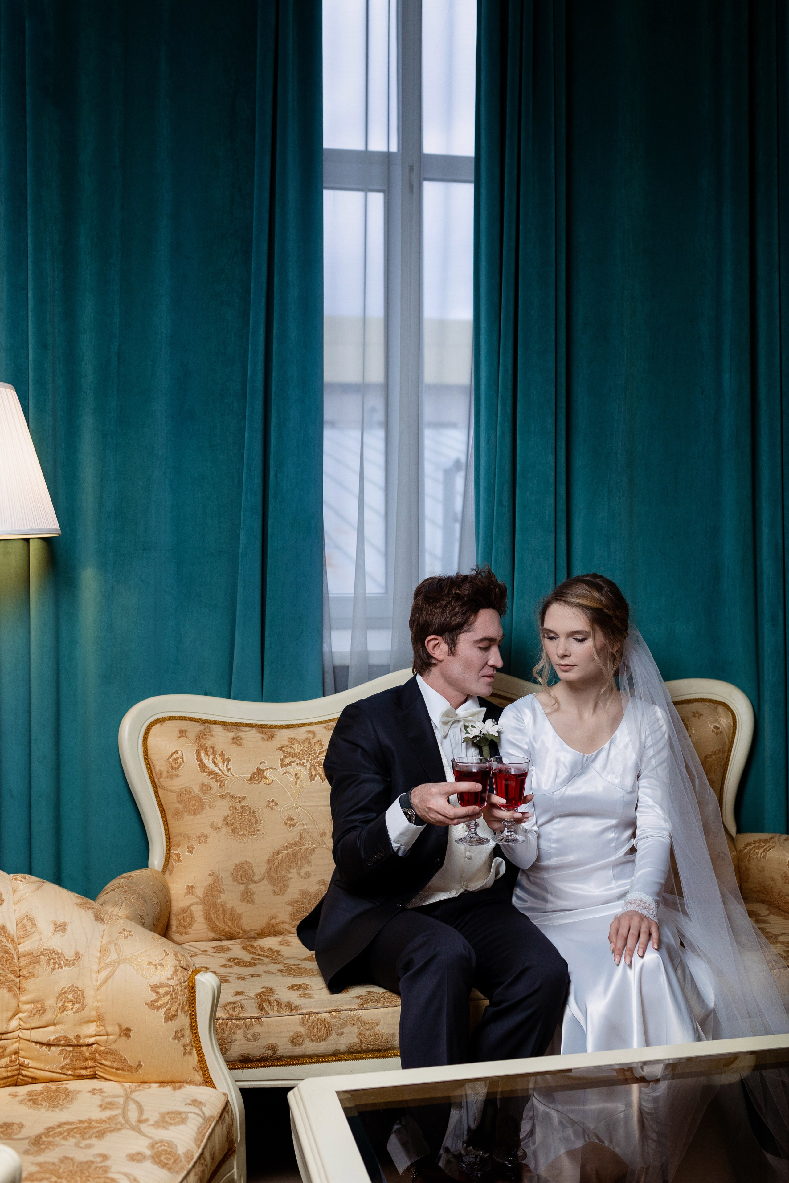 Жених с невестой сидят на диване с бокалами в руках на фоне зашторенного окна.