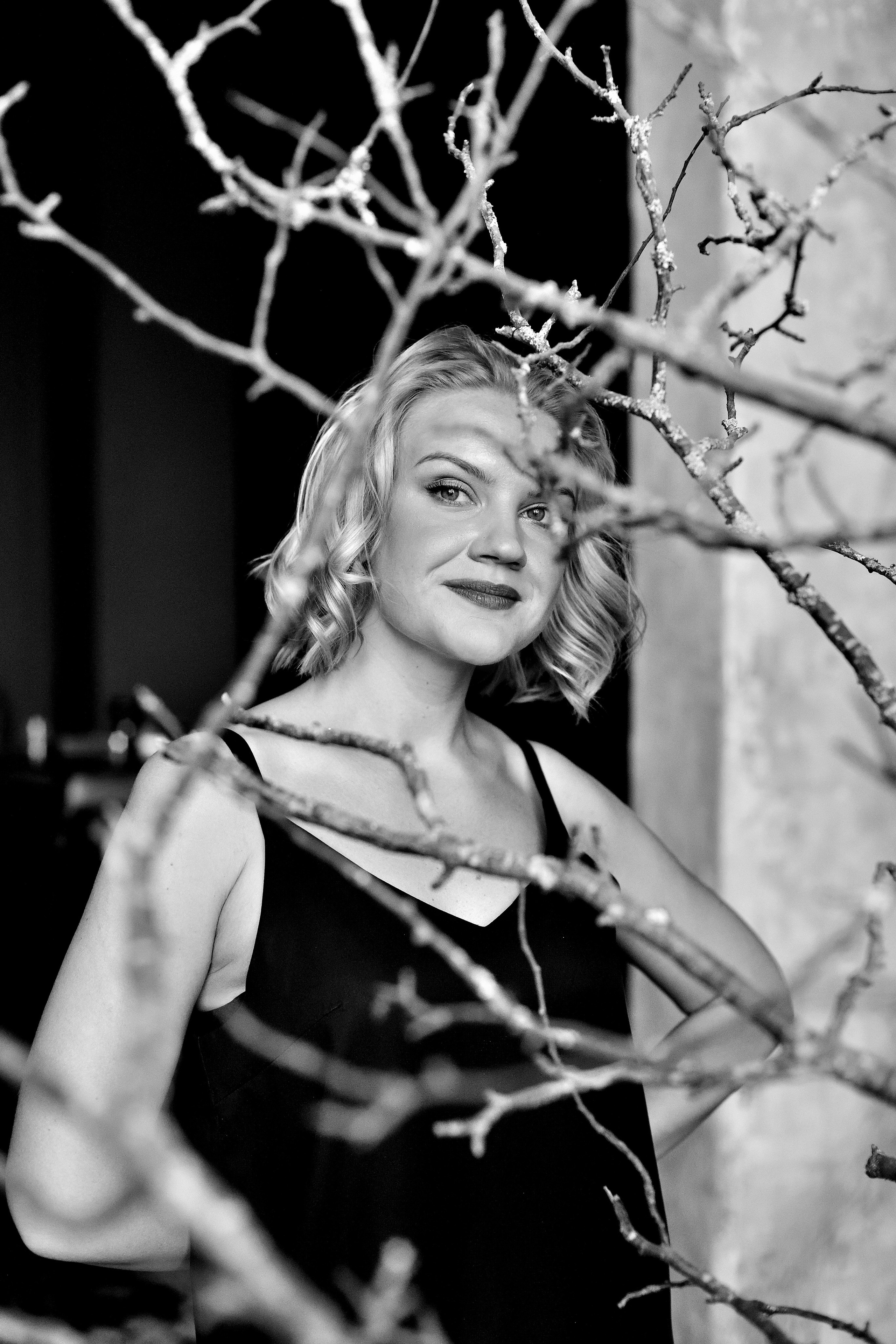 Девушка смотрит в объектив камеры через ветки деревьев.