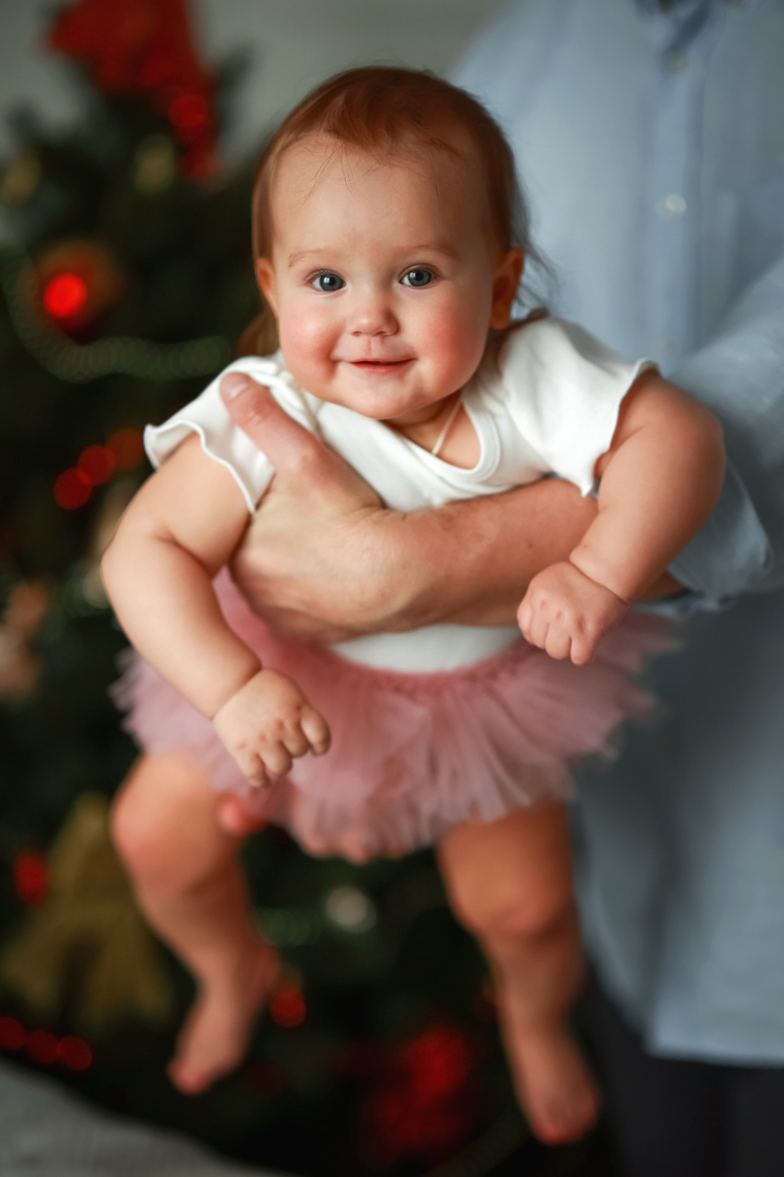 Улыбчивая малышка на папиных руках на фоне новогодней елки.