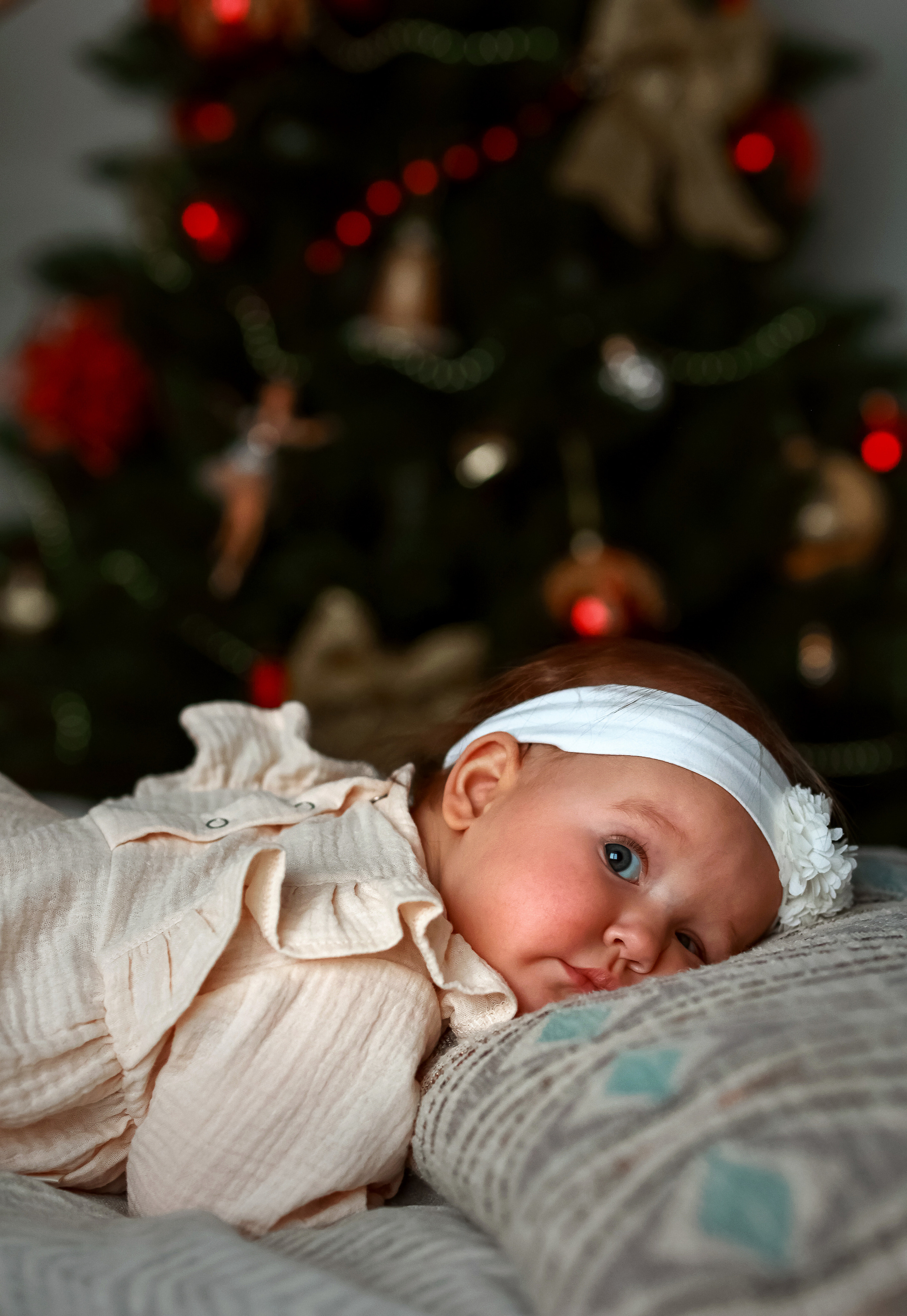 Маленькая девочка восьми месяцев с голубыми глазами задорно улыбается на фоне новогодней елки.