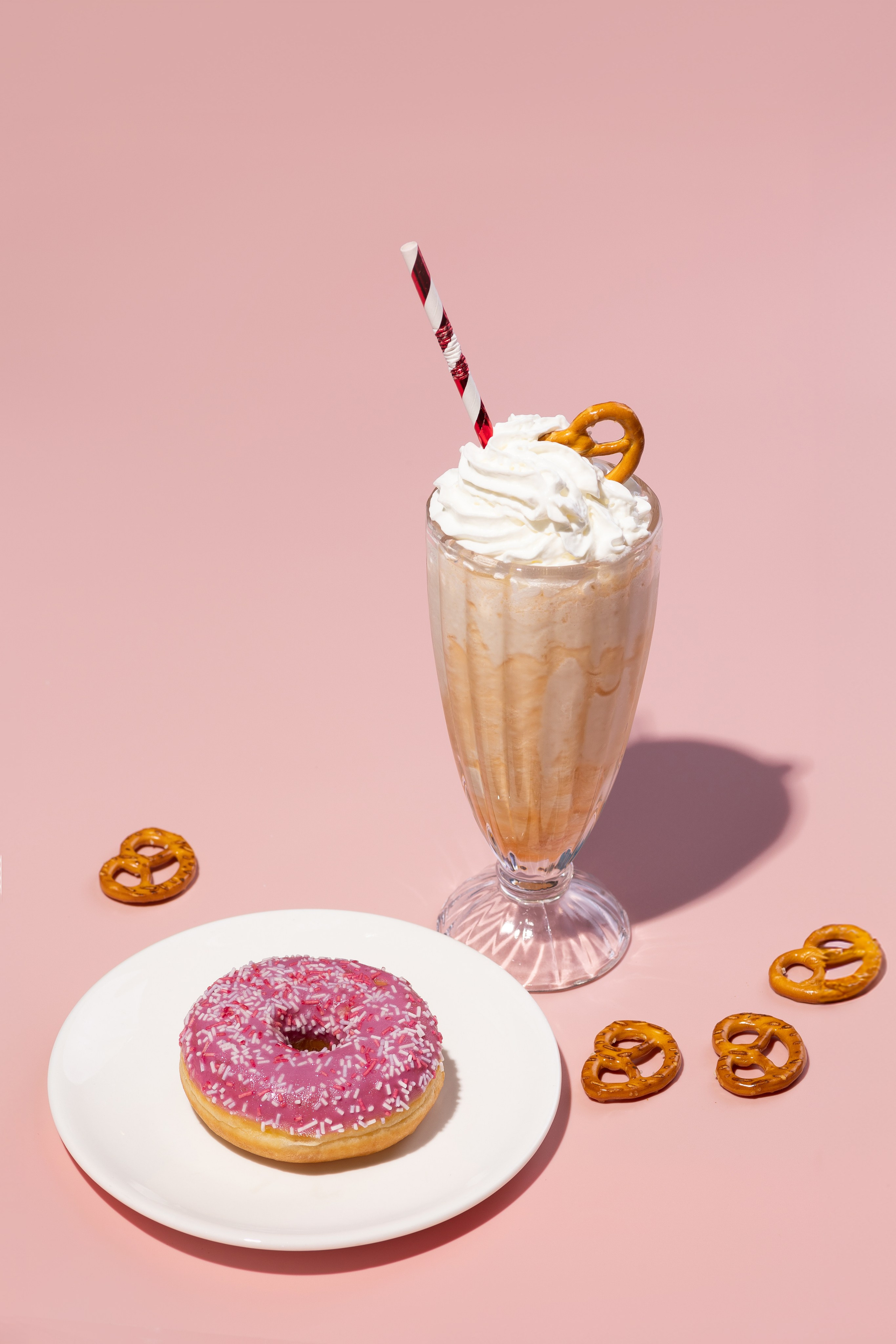 Молочный коктейль в стакане с трубочкой и пончик с розовой глазурью стоят на розовом фоне