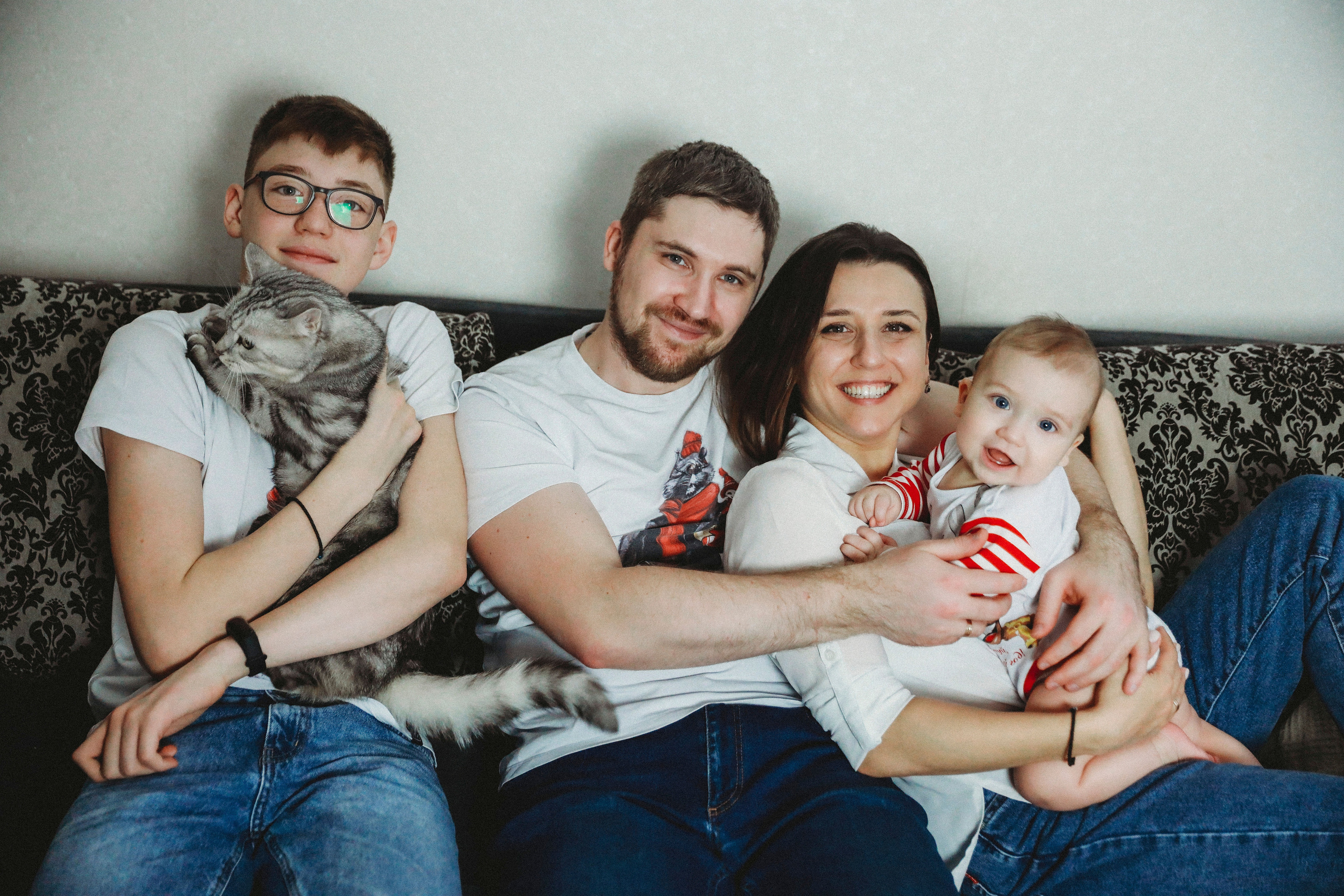 красивая молодая семья в белых  майках и джинсах сидит на диване у себя лома и держит котика у себя на руках