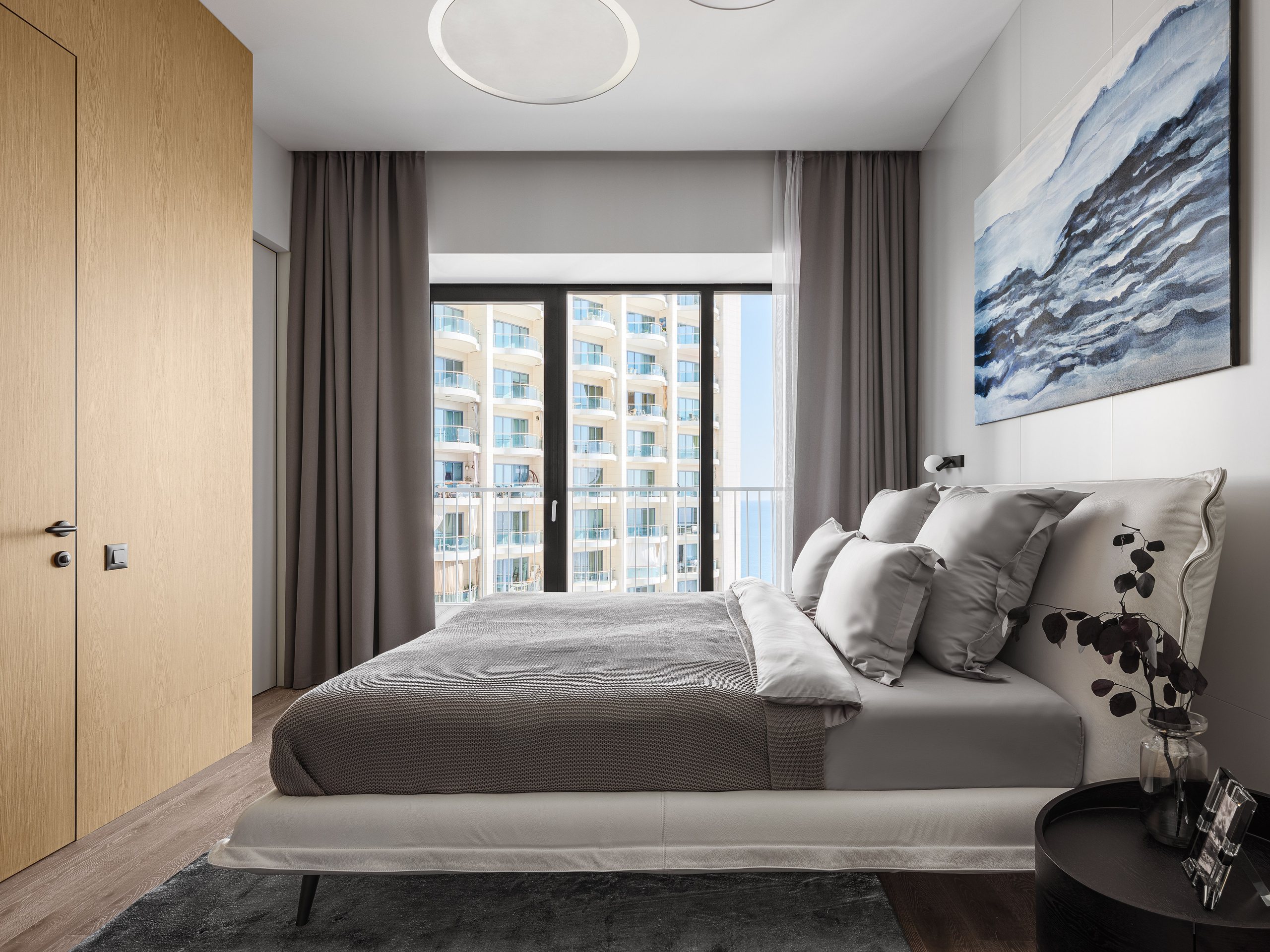 Стильная светлая спальня в ЖК Сочи с панорамным окном, серыми шторами и дизайнерской кроватью с текстилем в спокойной гамме. Авторский проект от Рената Байчукова и Алеcи Кириенко, стилизация Екатерины Ракитиной, фото Сергея Денисенко.
