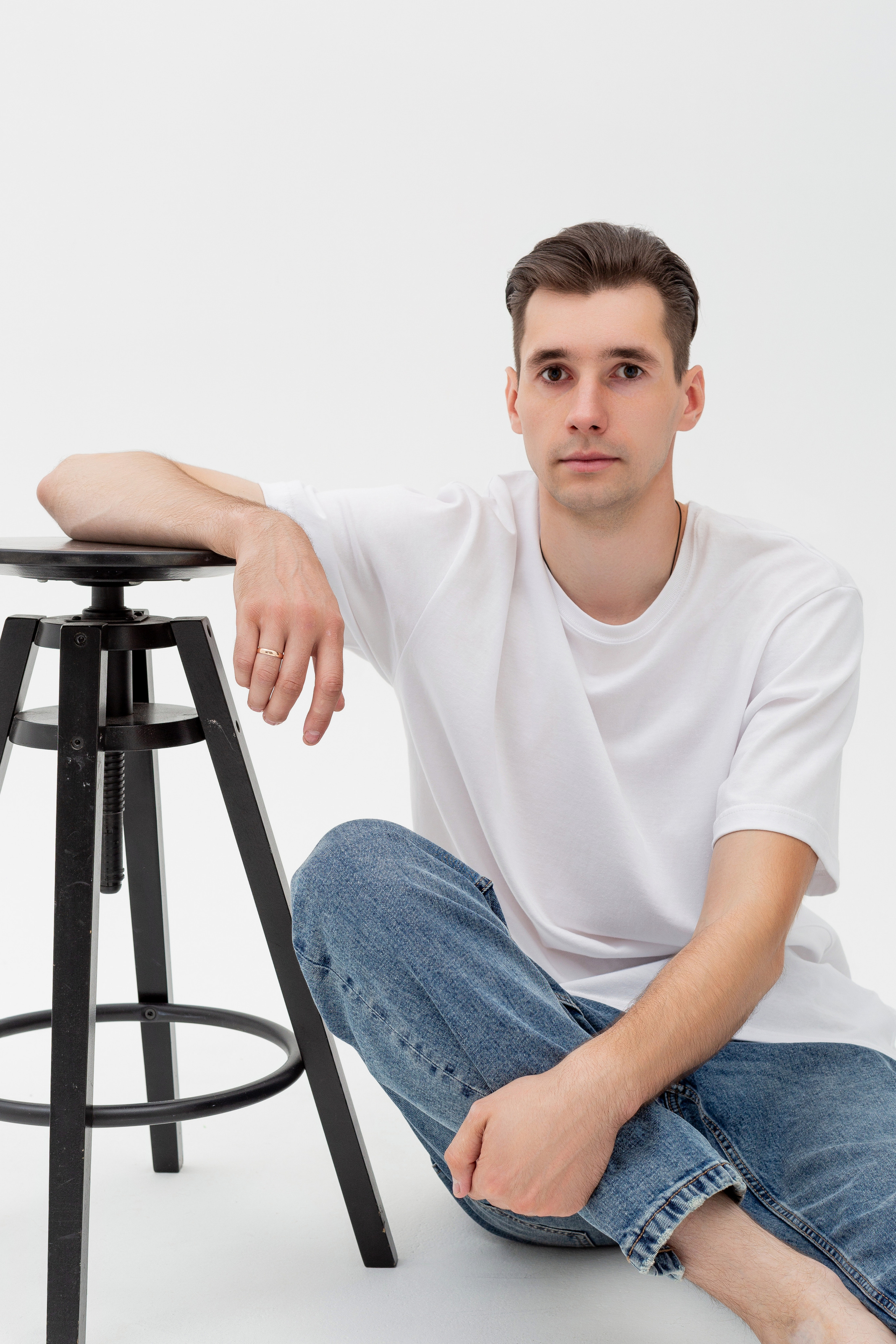 Студийный портрет мужчины сидящем около стула на полу, портрет 2/3, в белой футболке и джинсах на белом фоне