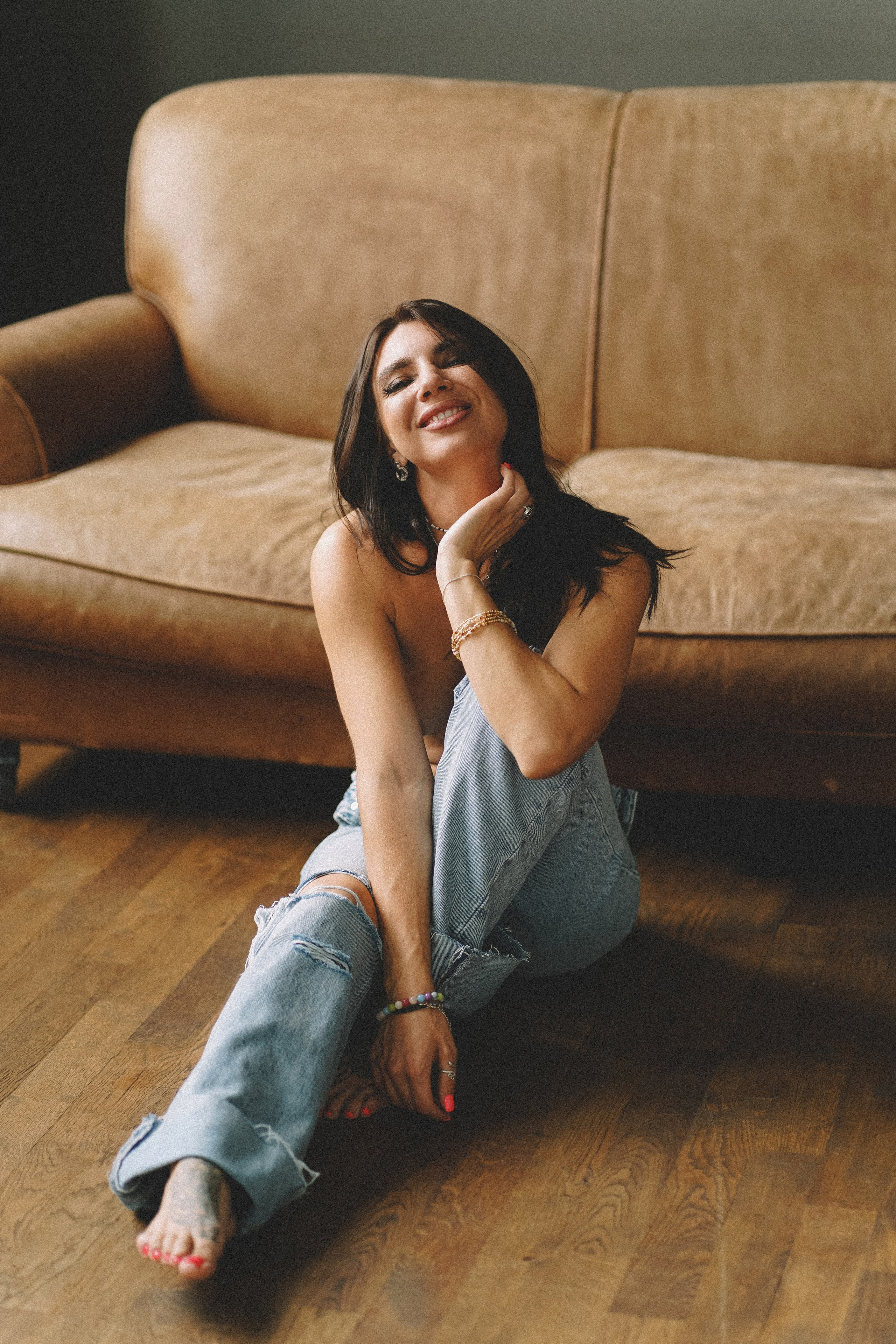 Девушка с темными волосами в джинсах сидит на полу около коричневого кожаного дивана и улыбается 