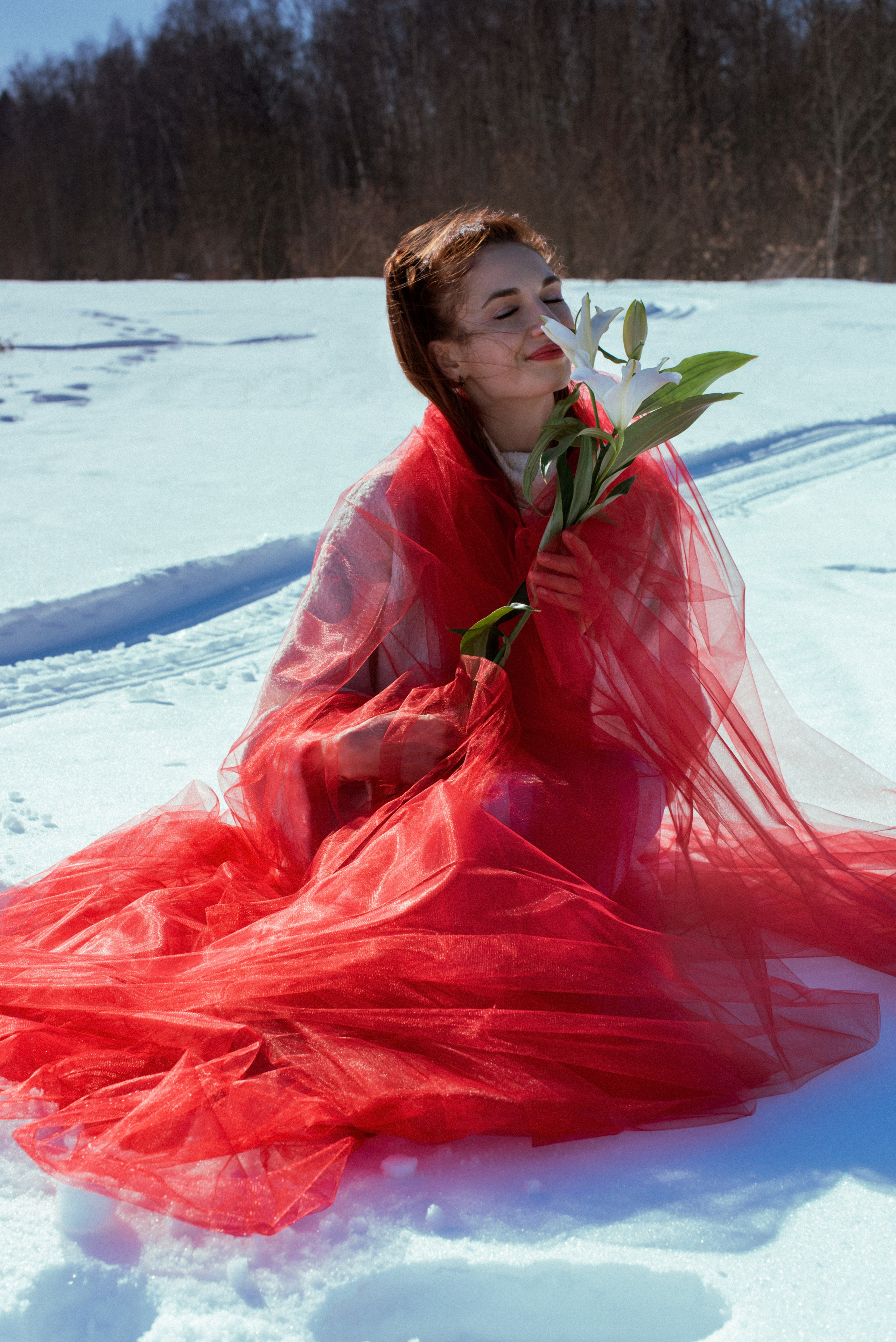 Девушка в белом свитере, укутанная в красный фатин, на фоне зимнего пейзажа.