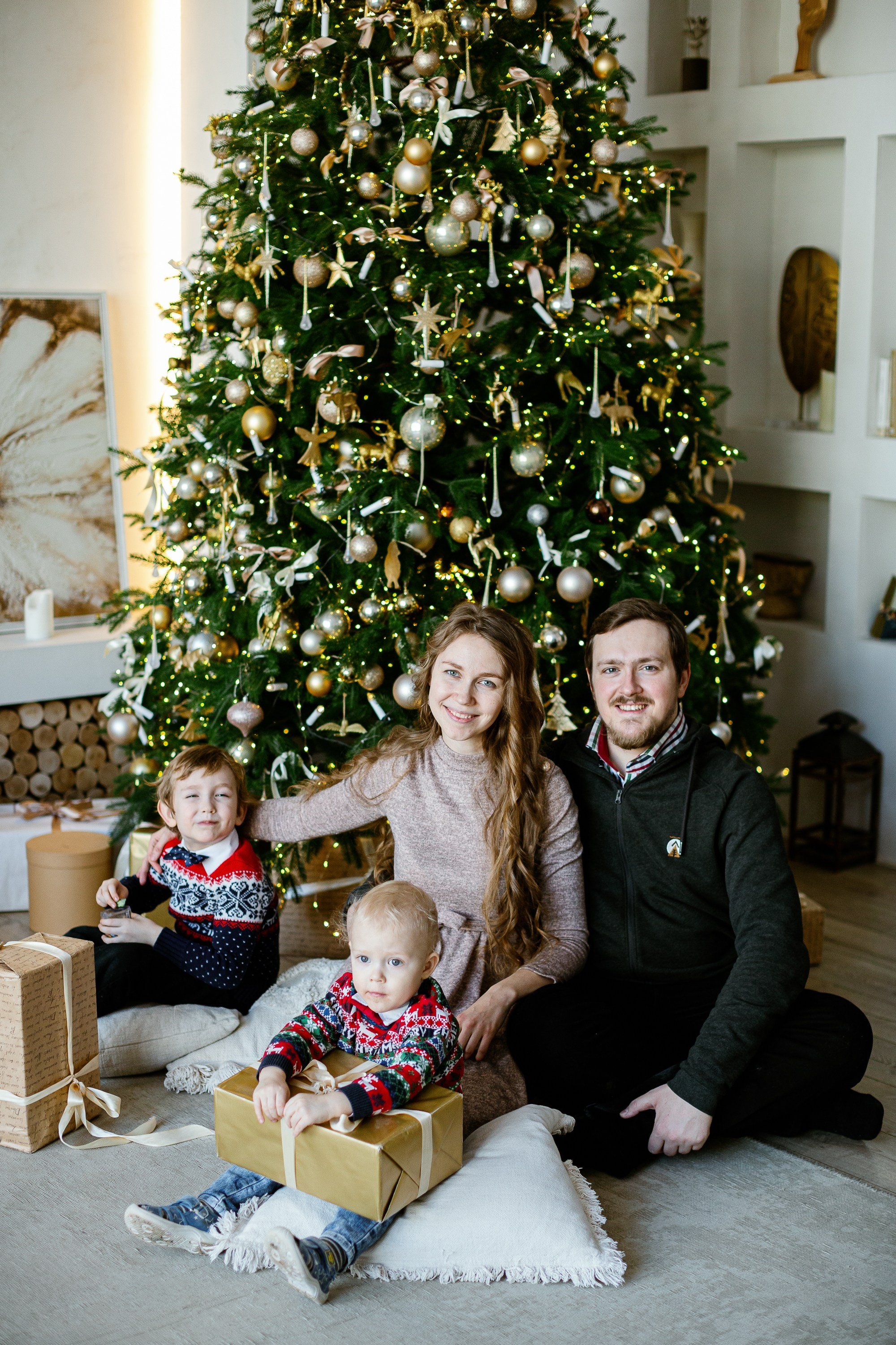 Семейная сидит на полу на фоне стоит елка и светится огоньками. Все улыбаются и смеются.